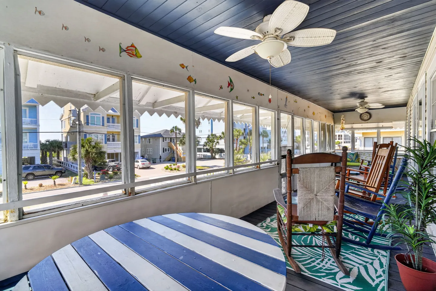 Screened porch with rocking chairs, round striped table, ceiling fans, and views of neighborhood houses.