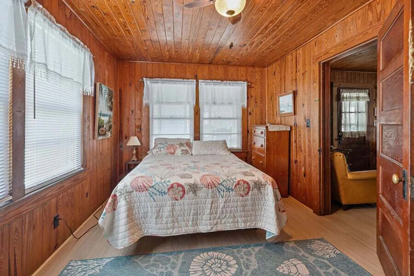 Bedroom with wood-paneled walls and ceiling, featuring a bed with a seashell-patterned quilt, windows with white blinds, and an area rug.