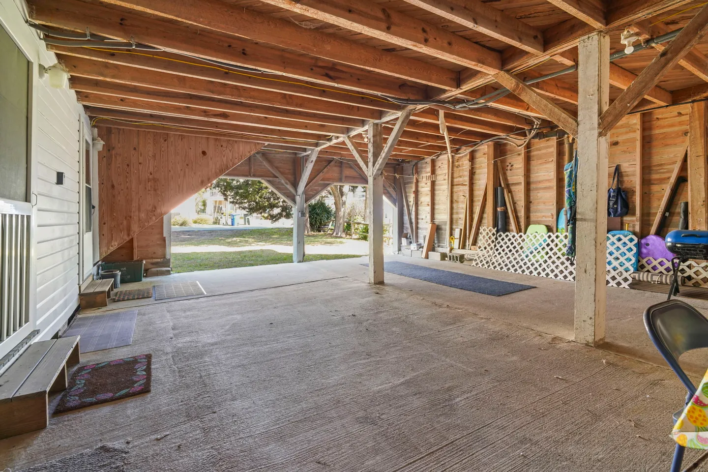 Open, covered ground-level storage and parking area beneath a raised house with concrete floor and exposed wooden beams.