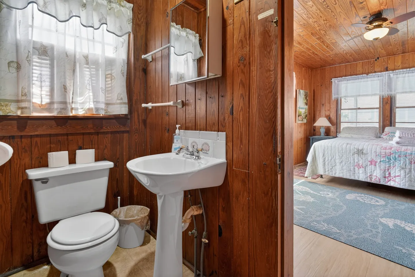 Bathroom with wood-paneled walls, pedestal sink, and toilet, looking into an adjoining bedroom.