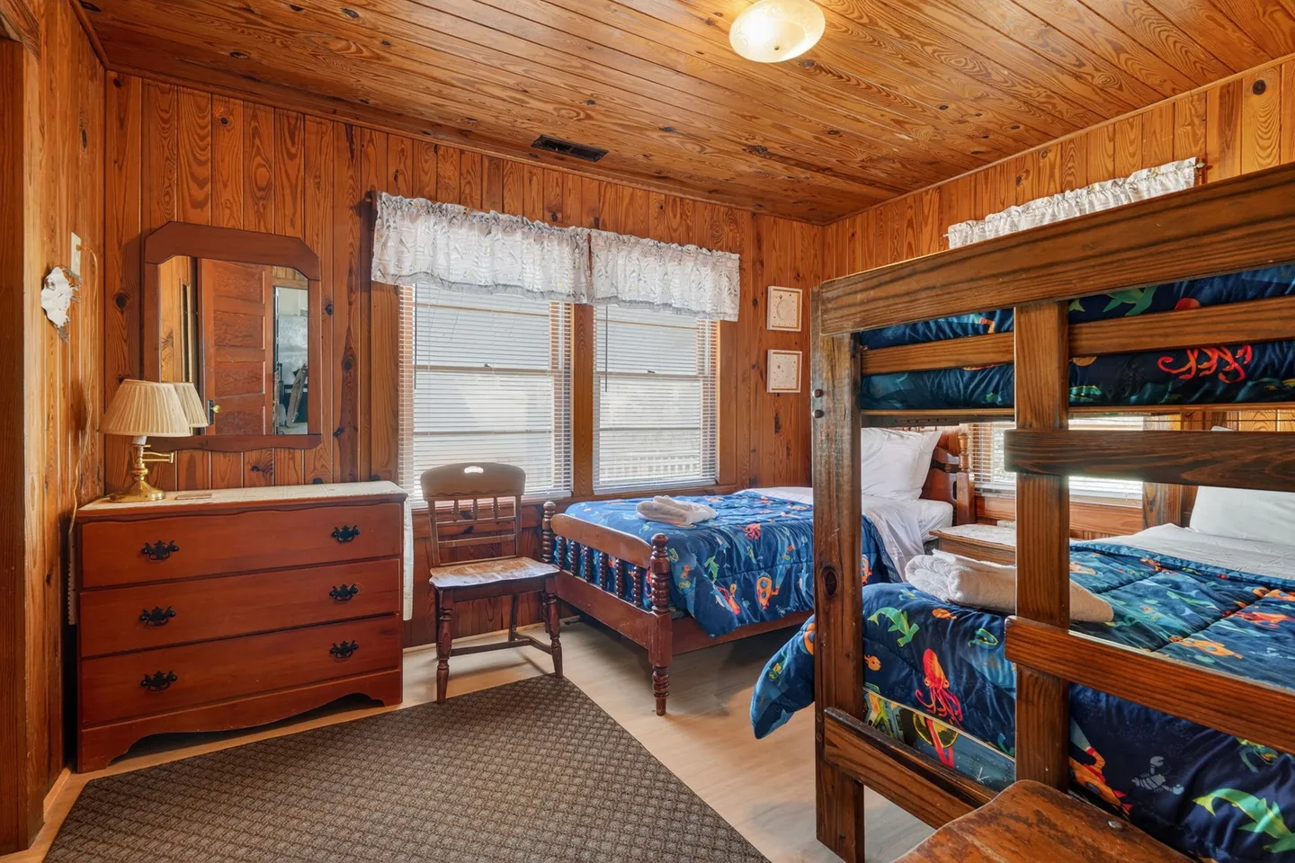 Bedroom with wood-paneled walls and ceiling, featuring wooden bunk beds, a single bed with colorful ocean-themed bedding, a dresser, and windows with blinds.