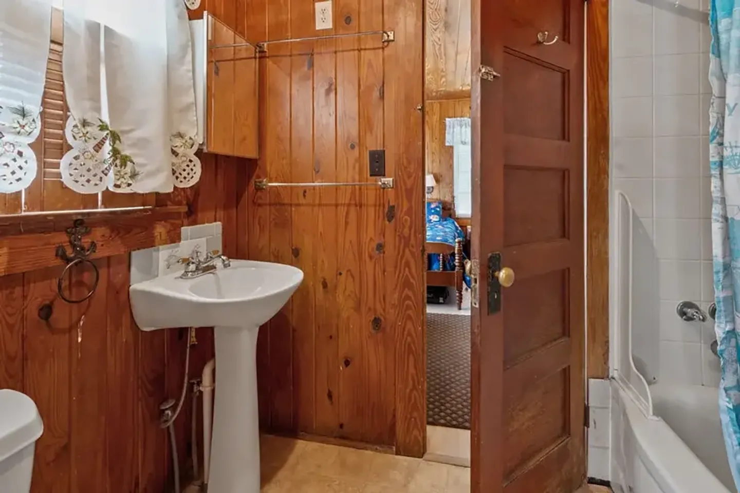 Bathroom with wood-paneled walls, pedestal sink, towel bars, and a view into an adjacent bedroom.