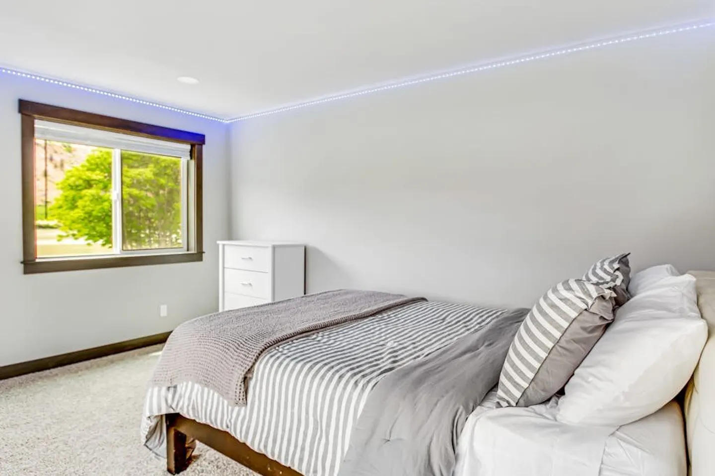 Bedroom with a bed dressed in striped and grey linens, a window with outdoor views, light carpet, and LED strip lighting near the ceiling.