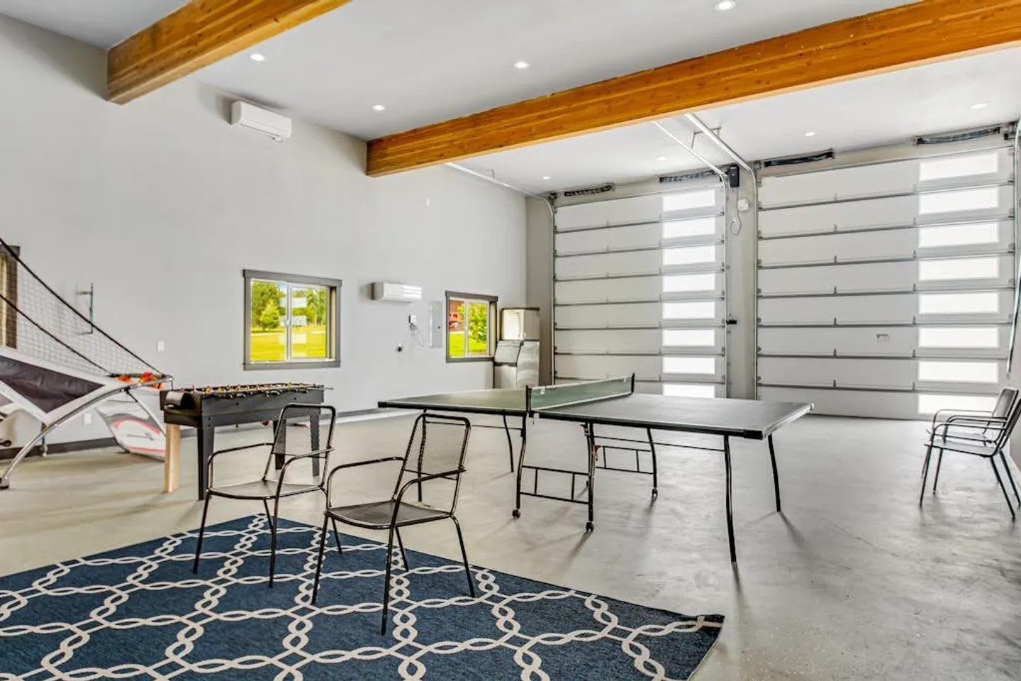 Spacious garage converted into a game room with exposed ceiling beams, a ping pong table, foosball, and a basketball arcade game.