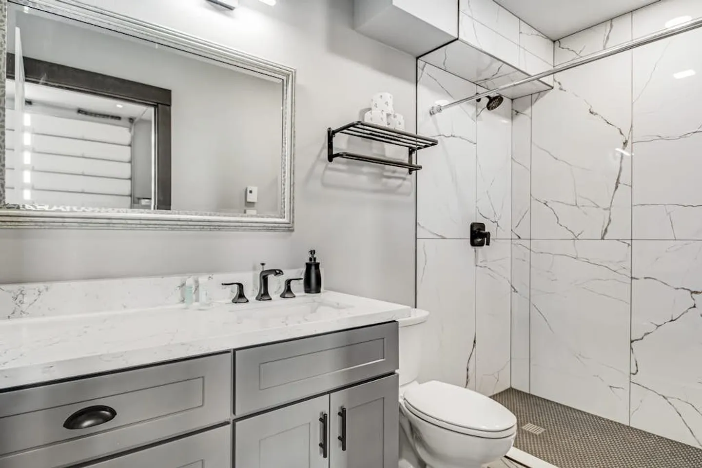 Bathroom with marble-style tiled walk-in shower, grey vanity with white countertop, framed mirror, and toilet.
