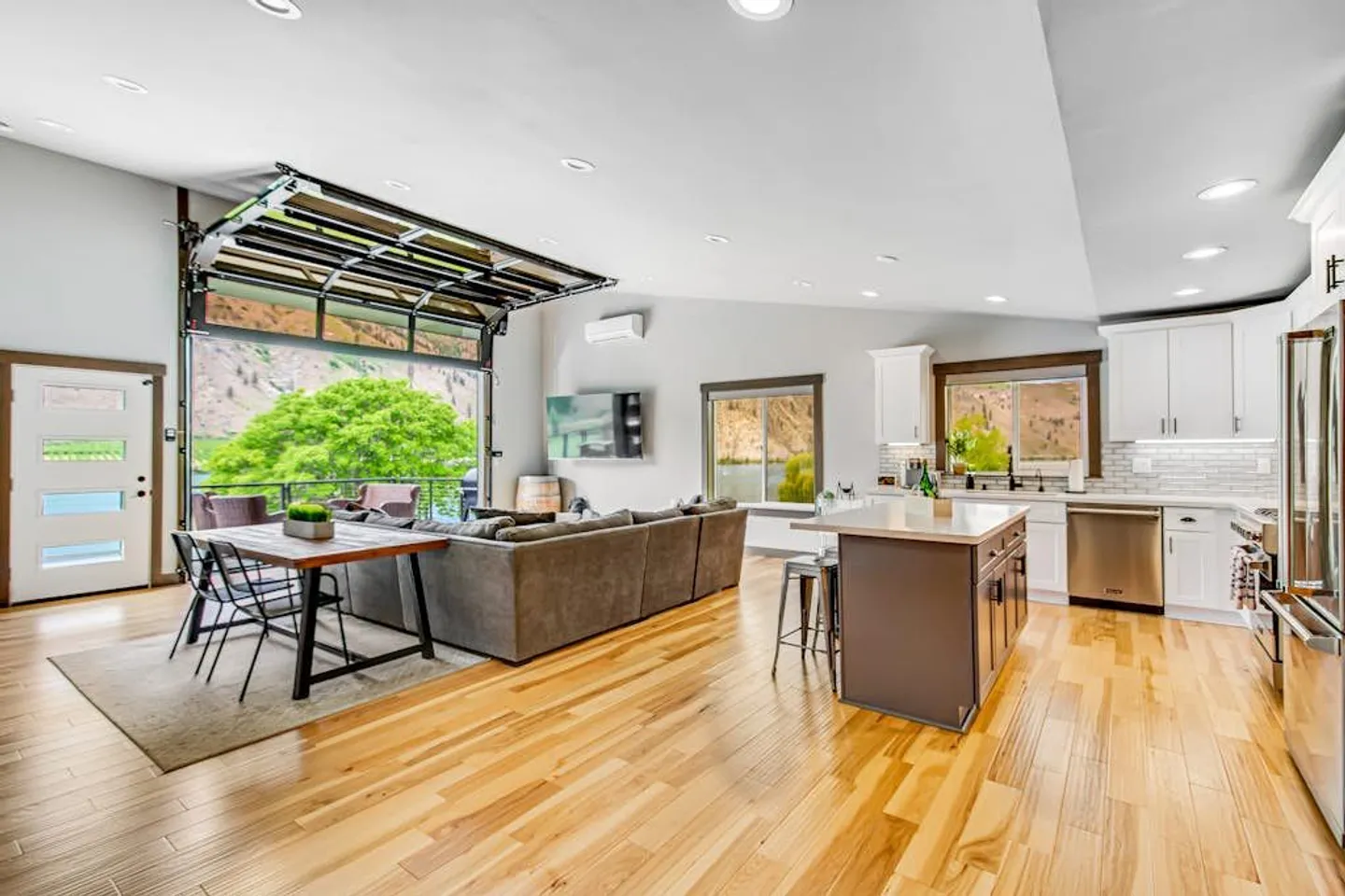 Open-concept living room, dining area, and kitchen featuring light wood floors and a raised roll-up glass garage door opening to a deck.