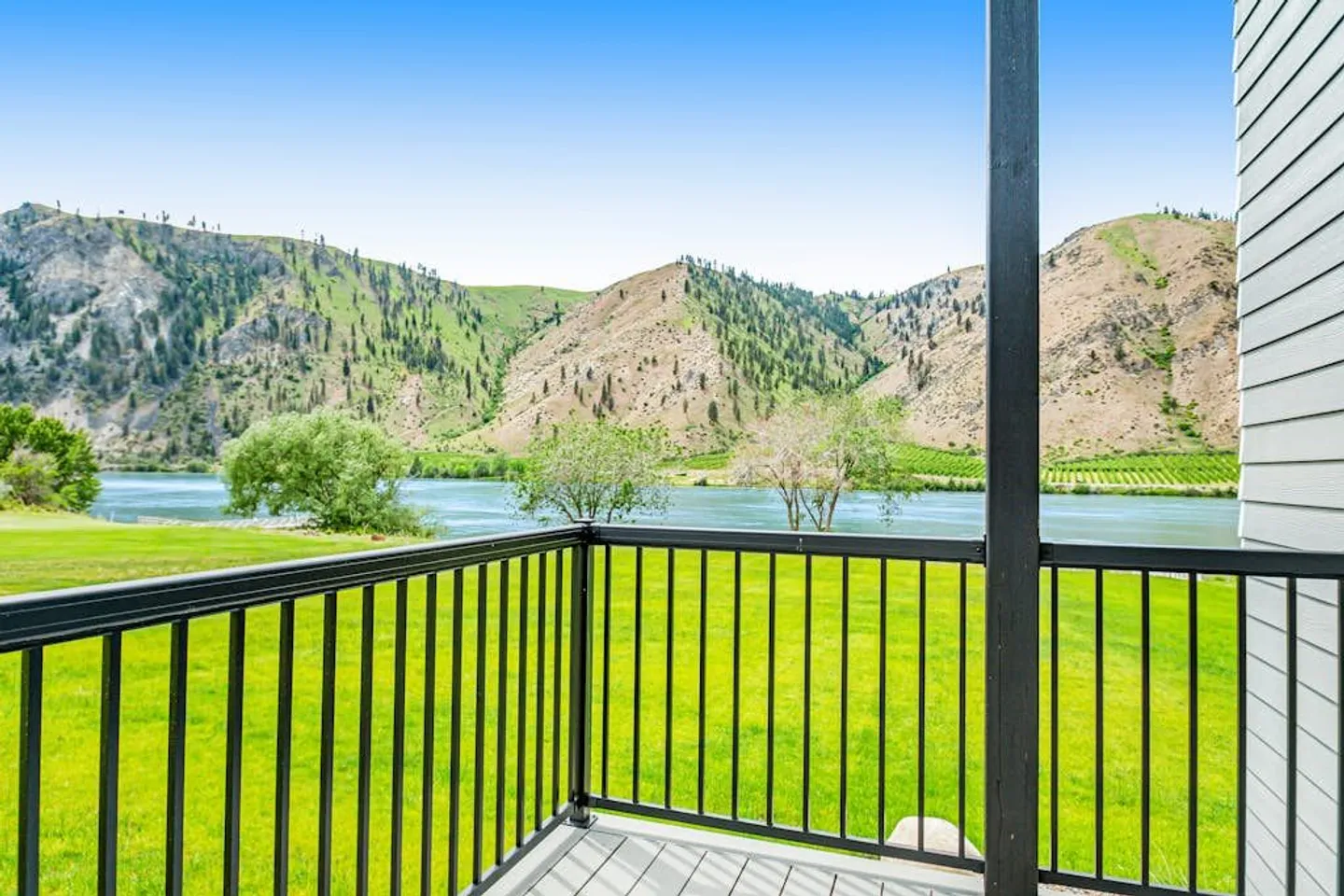View from an elevated deck with black railing overlooking a green lawn, a river, and hills under a blue sky.