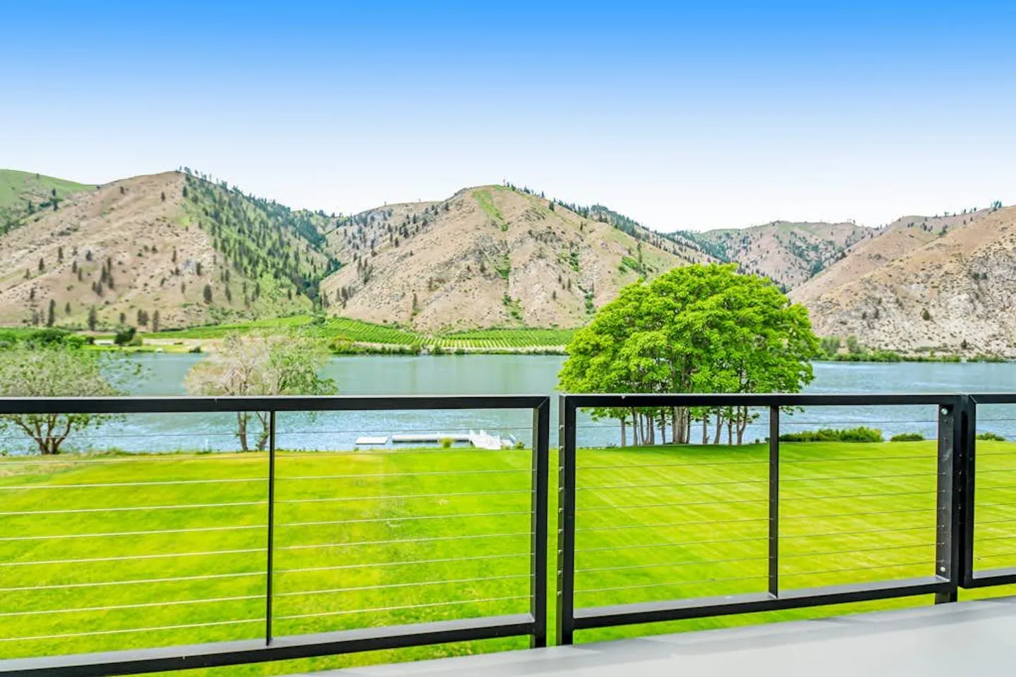 View from an elevated deck with black railing overlooking a green lawn, a river, and hills in the background.