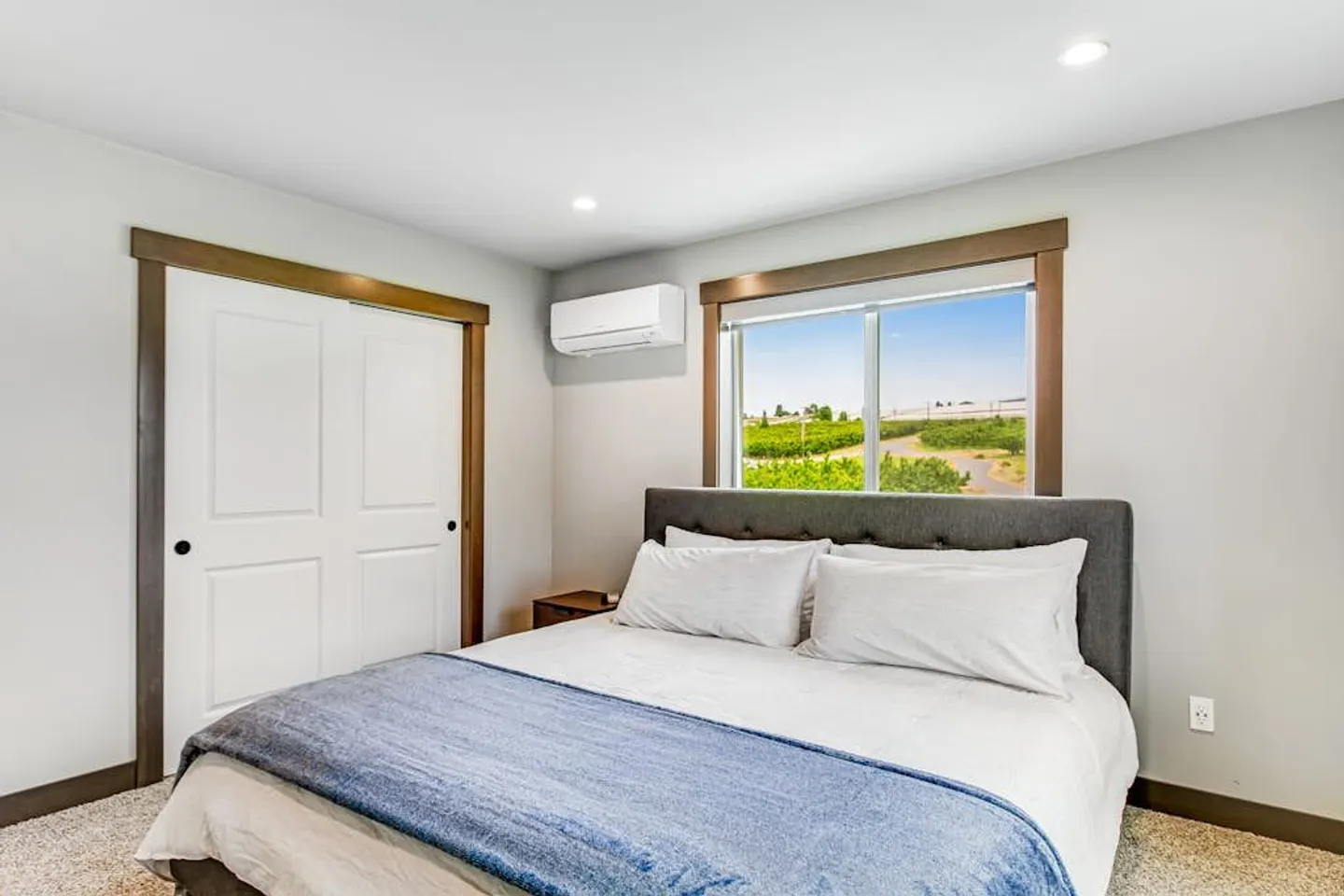 Bedroom with a bed dressed in white and blue bedding, sliding closet doors, wall-mounted air conditioner, and window looking out to fields.