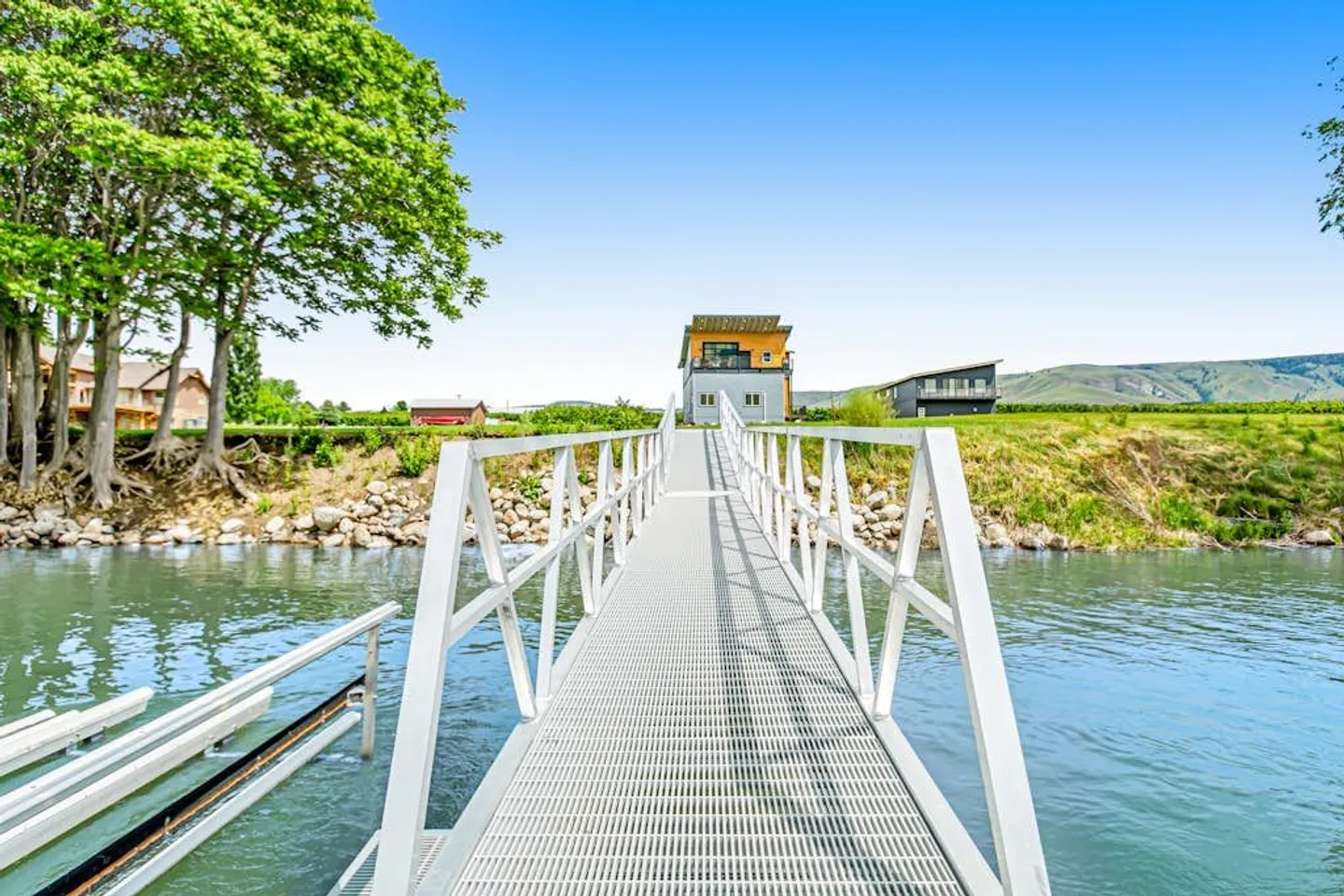 A metal gangway dock leading from the water toward a grassy shoreline and a two-story home under a clear blue sky.