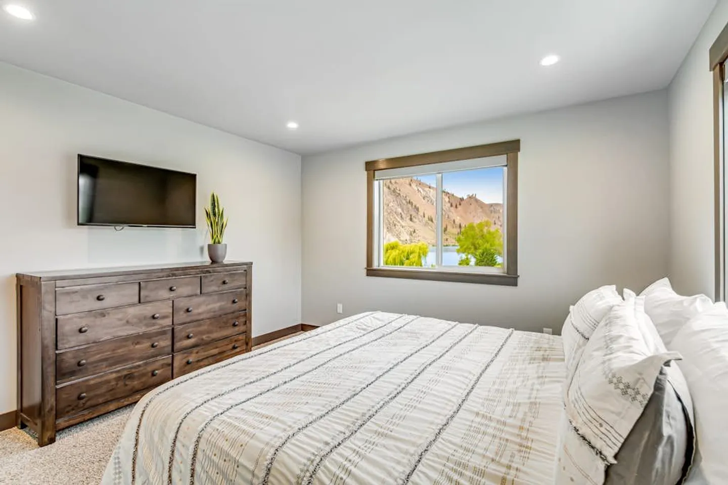Bedroom featuring a made bed with white patterned bedding, a dark wood dresser with a wall-mounted TV above, and a window with scenic outdoor views.