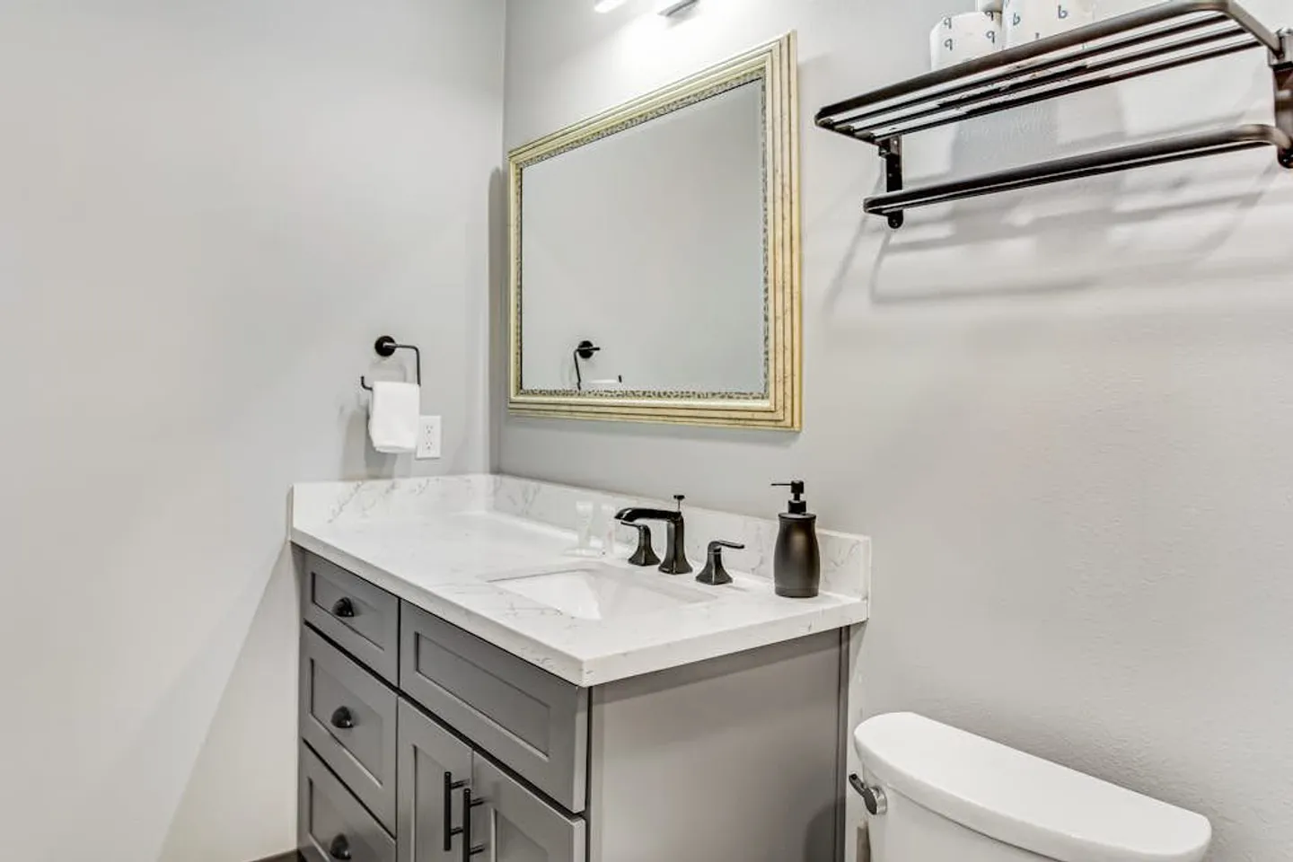 Bathroom vanity with a gray cabinet, marble-patterned countertop, framed mirror, and part of a toilet visible.