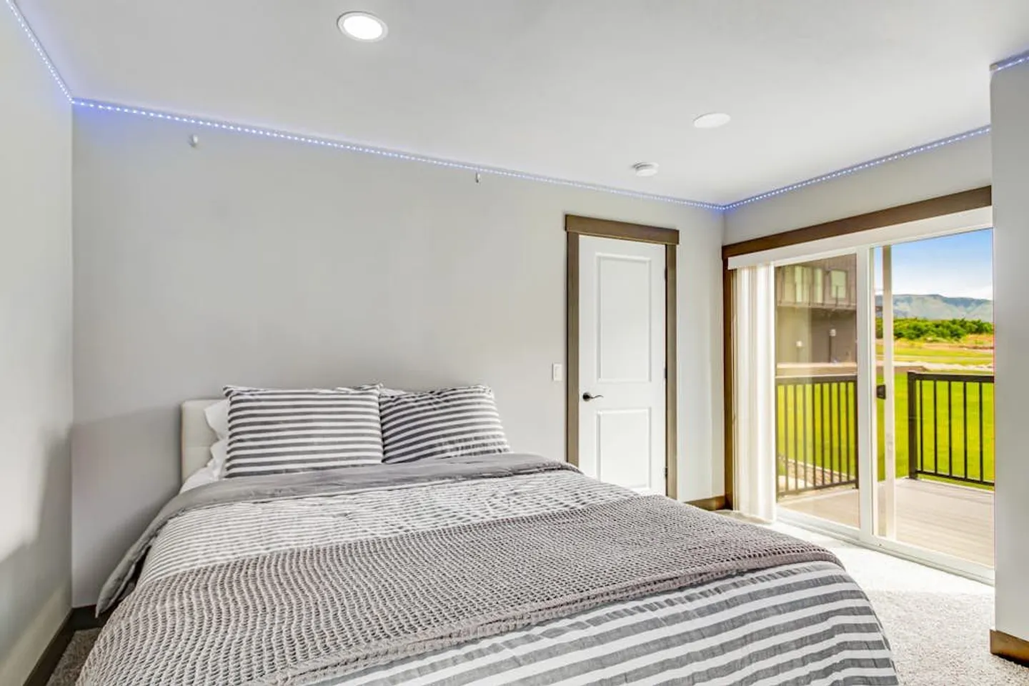 Bedroom with a bed made up with striped bedding, recessed ceiling lights with blue LED accent strip, and sliding glass doors leading to an outdoor balcony.