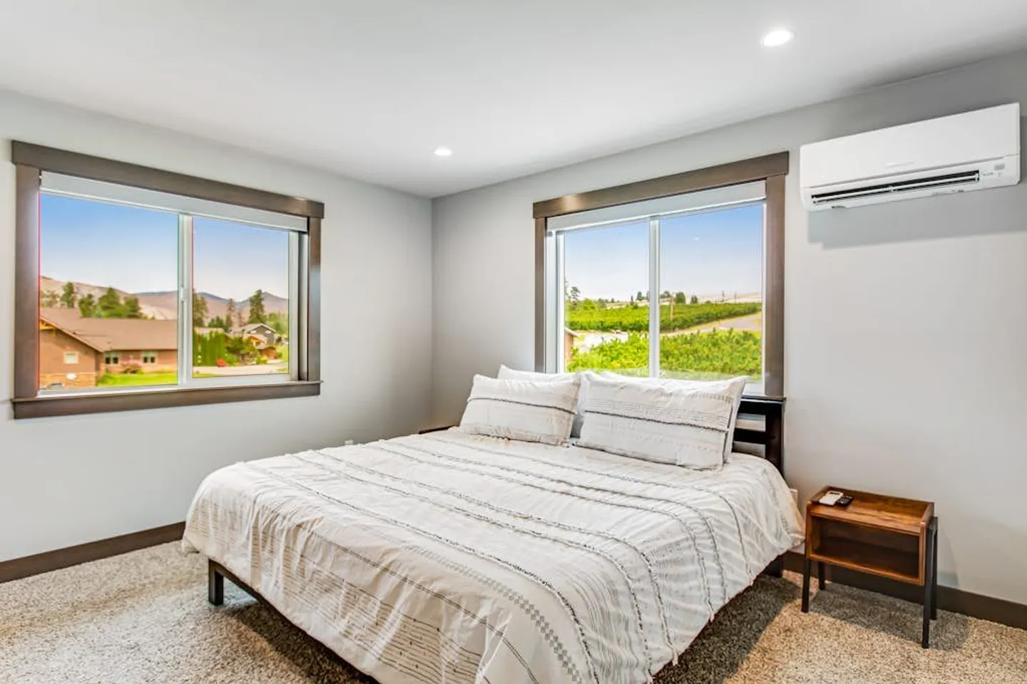 Bedroom with a made bed, two windows overlooking hills and an orchard, a nightstand, and a wall-mounted air conditioner.