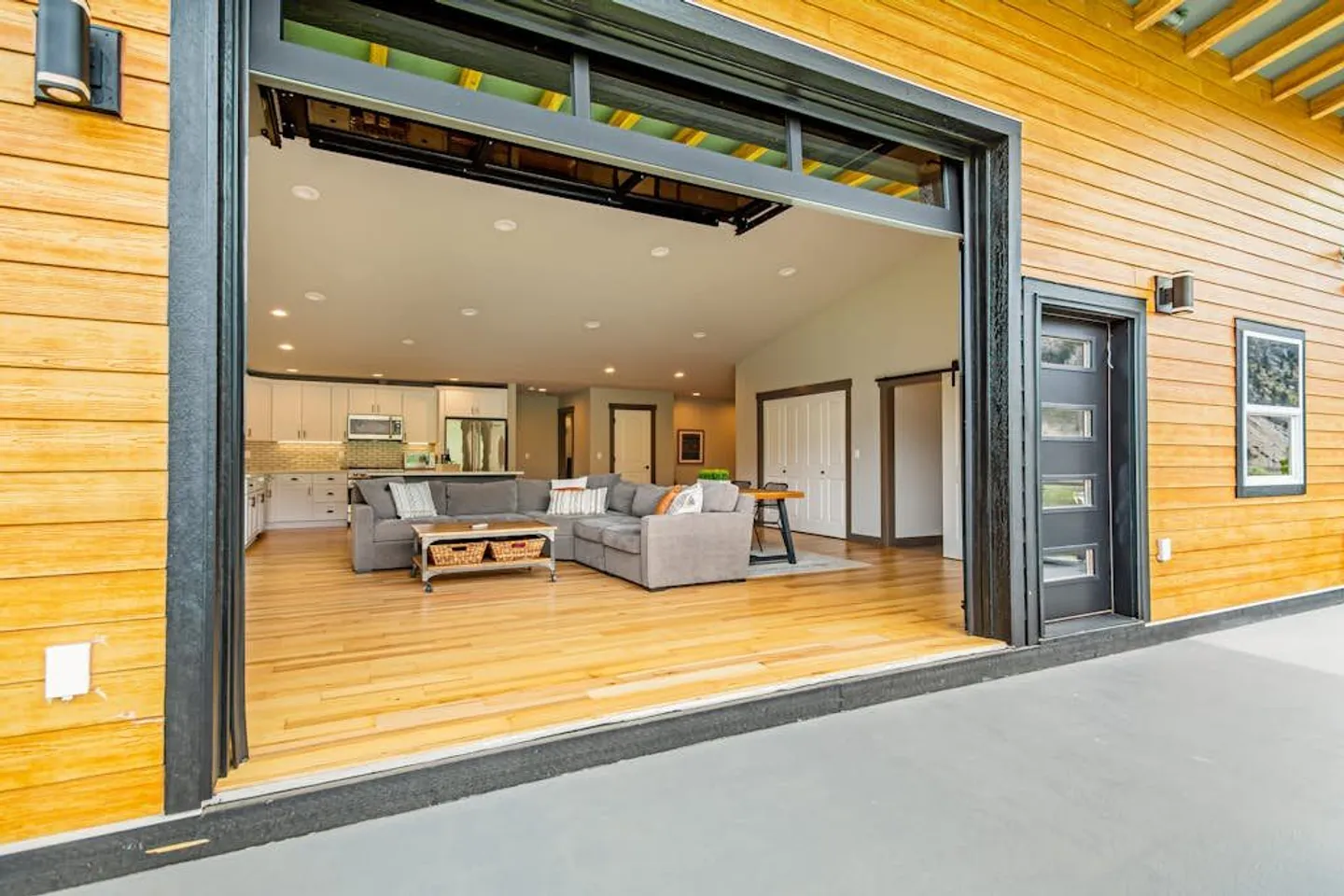 A wide roll-up glass garage door opens from an outdoor patio into an open-concept living room and kitchen with wood floors and a gray sectional sofa.