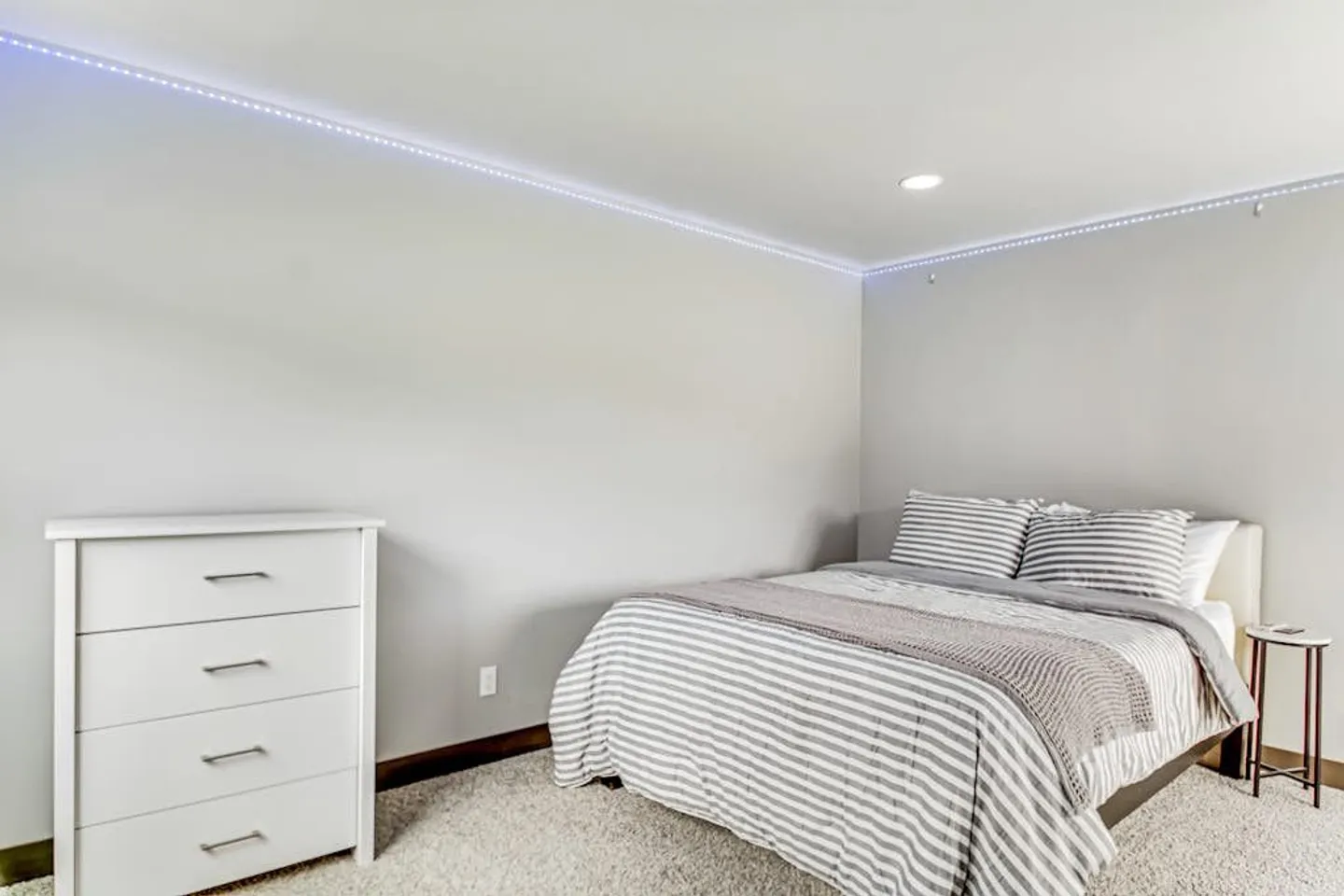 Bedroom with carpet, striped bedding, a white four-drawer dresser, a small nightstand, and LED strip lighting along the ceiling edge.
