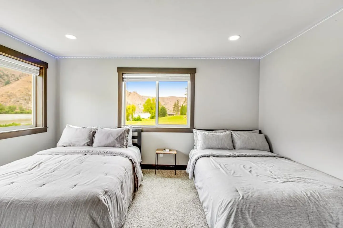 Bedroom with two beds made up in light gray bedding, light gray walls, carpeted flooring, and windows overlooking hills outside.