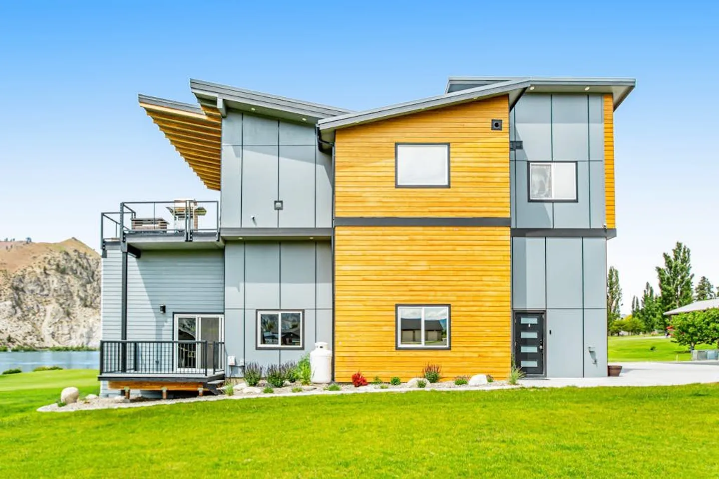 Exterior of a modern two-story home featuring grey paneling, wood siding, balconies, and a green lawn under a bright blue sky.