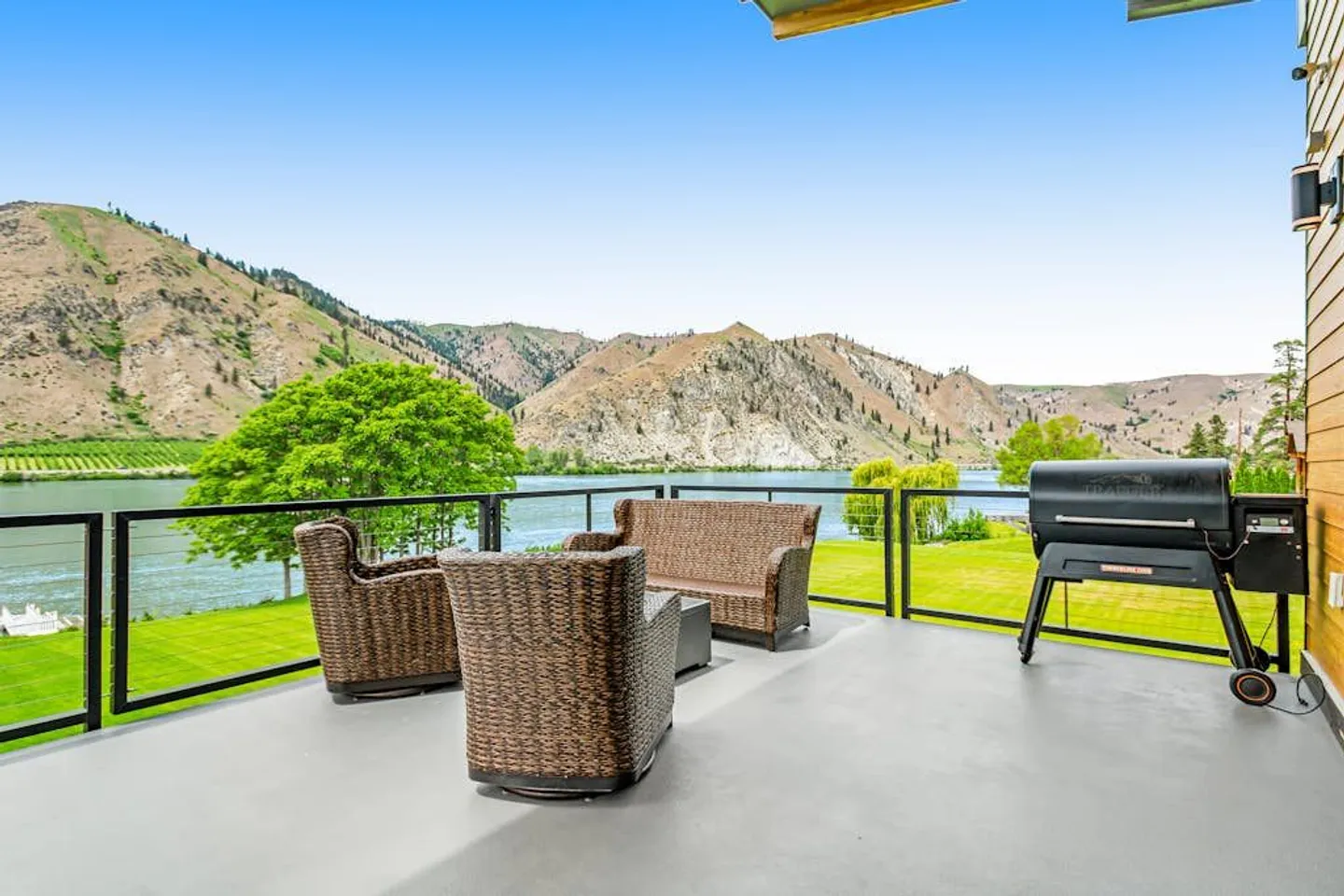 Outdoor deck with woven wicker seating, a pellet grill, and railings overlooking a lake and surrounding mountains.