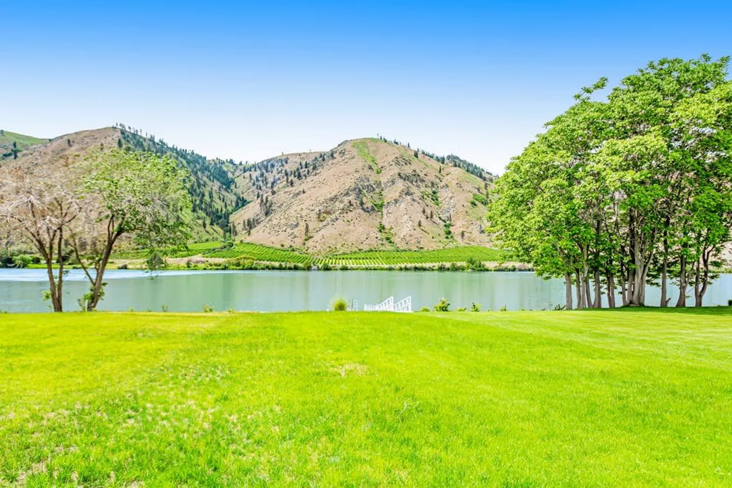 A wide view of a green lawn leading down to a river, with arid mountains and a clear blue sky across the water.
