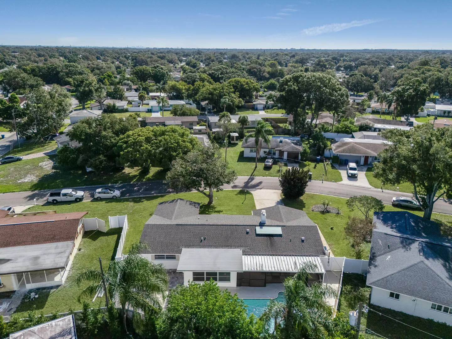 Aerial view of a single-story house with a backyard swimming pool surrounded by a residential neighborhood with green trees under a blue sky.
