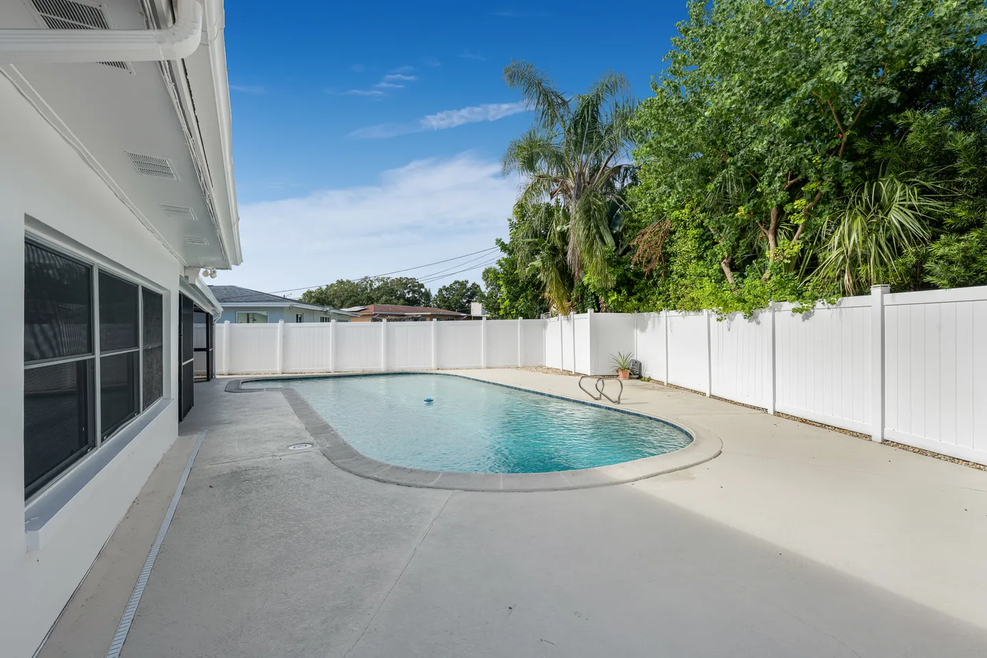 Outdoor swimming pool in a paved backyard bordered by a white privacy fence and palm trees under a blue sky.