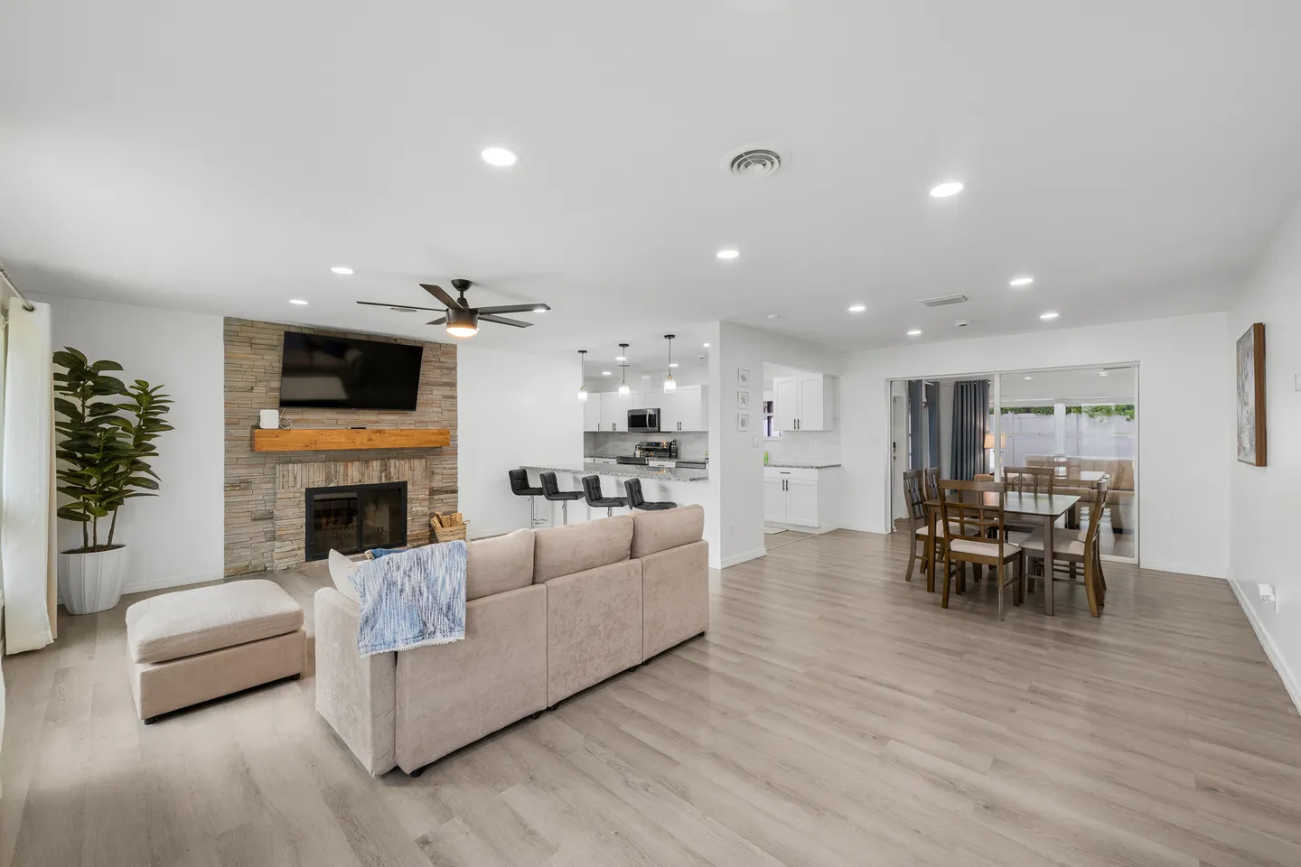 Open-concept living room, kitchen, and dining area featuring light wood-look floors, a stone fireplace with a mounted TV, and recessed lighting.