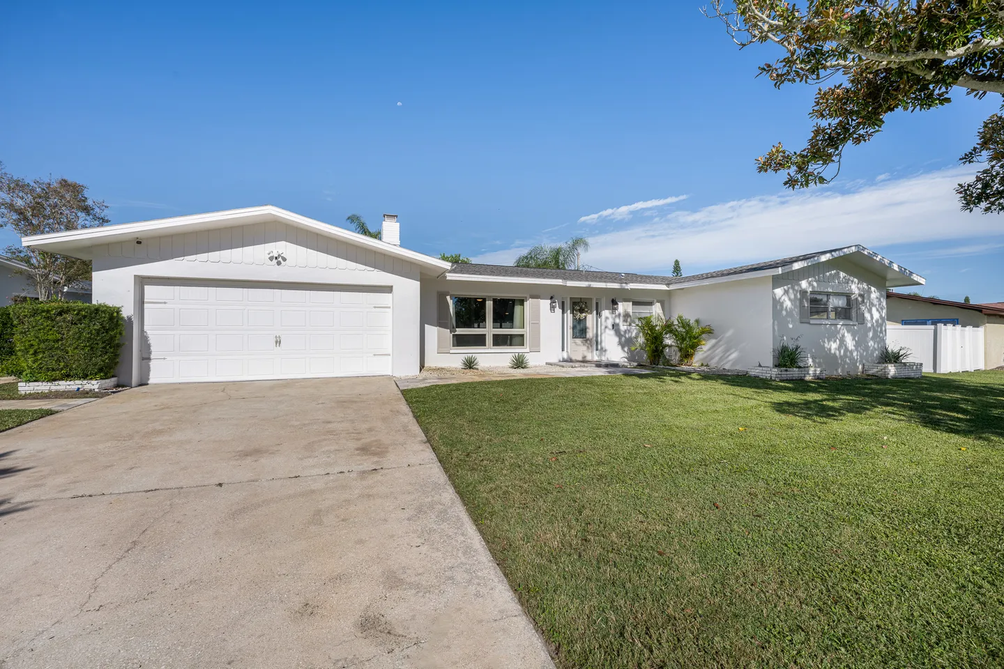 Front exterior of a single-story white house with a two-car garage, concrete driveway, and green lawn under a blue sky.