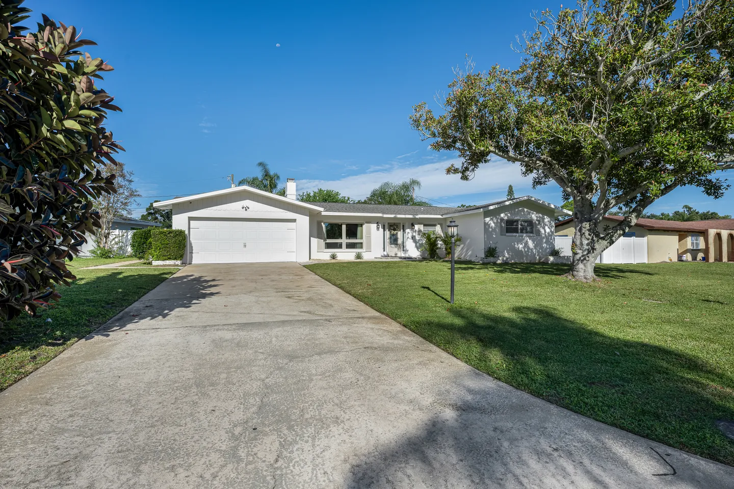 Front exterior of a single-story white home with a long concrete driveway, front lawn, and mature trees under a clear blue sky.