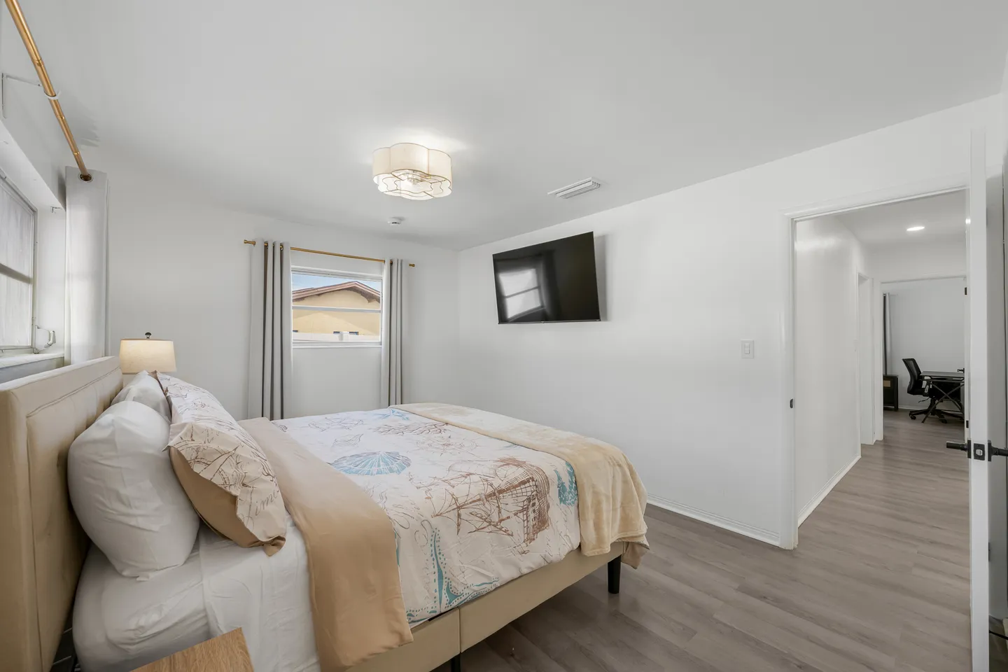 Bedroom with a made bed, wall-mounted television, wood-look flooring, and an open doorway leading to a hallway.