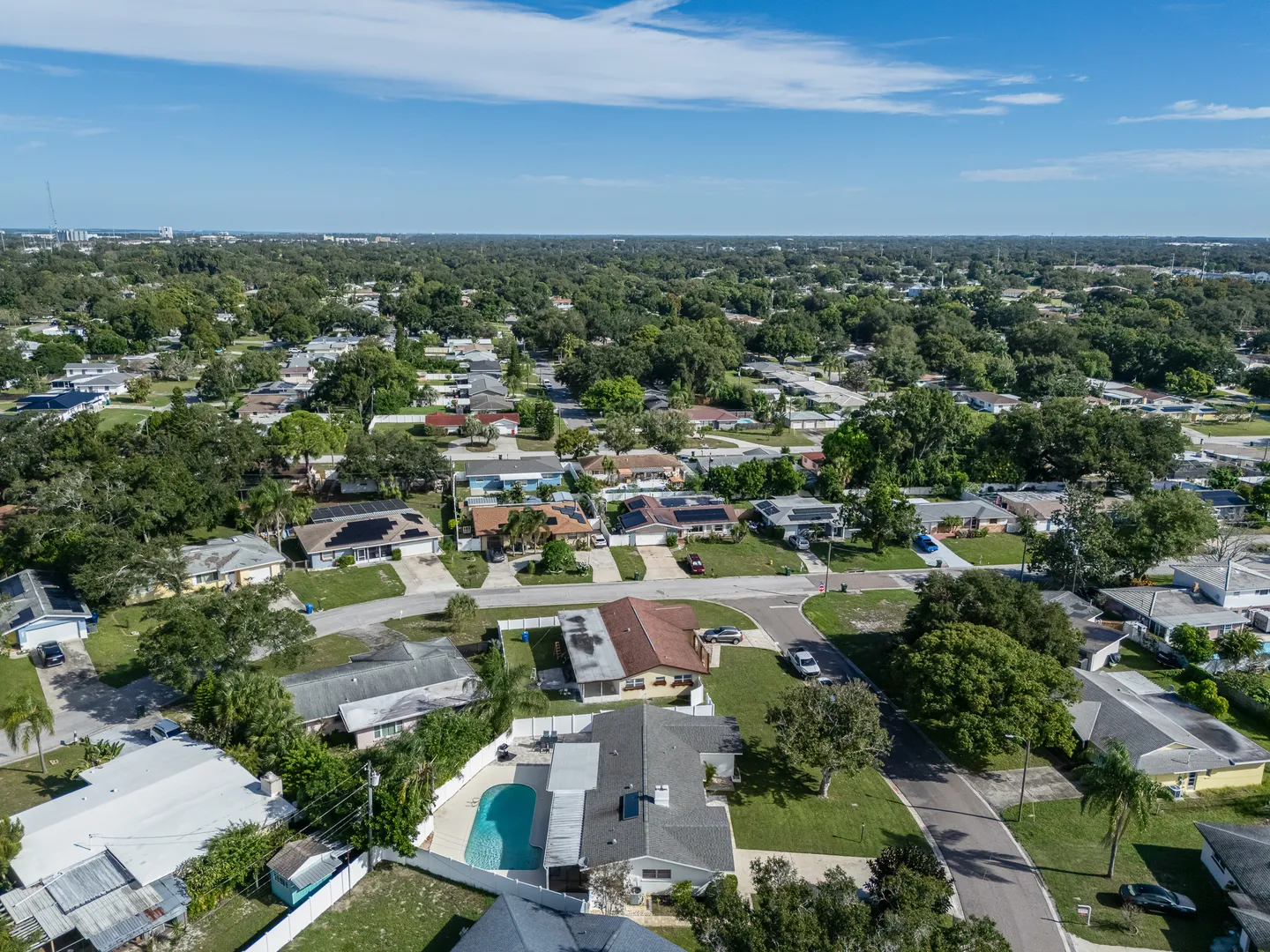 High-angle aerial view of a suburban neighborhood featuring single-story houses, lush green trees, paved streets, and a backyard swimming pool under a blue sky.