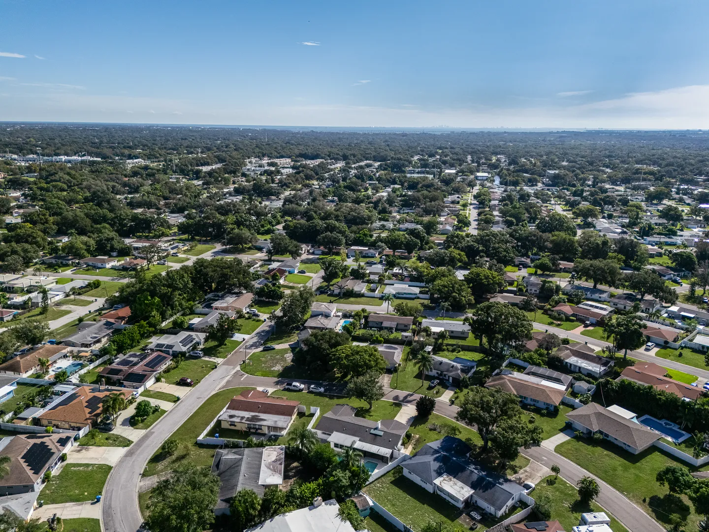 High-angle aerial photograph of a residential suburban neighborhood with single-story homes, green lawns, mature trees, and paved streets under a blue sky.