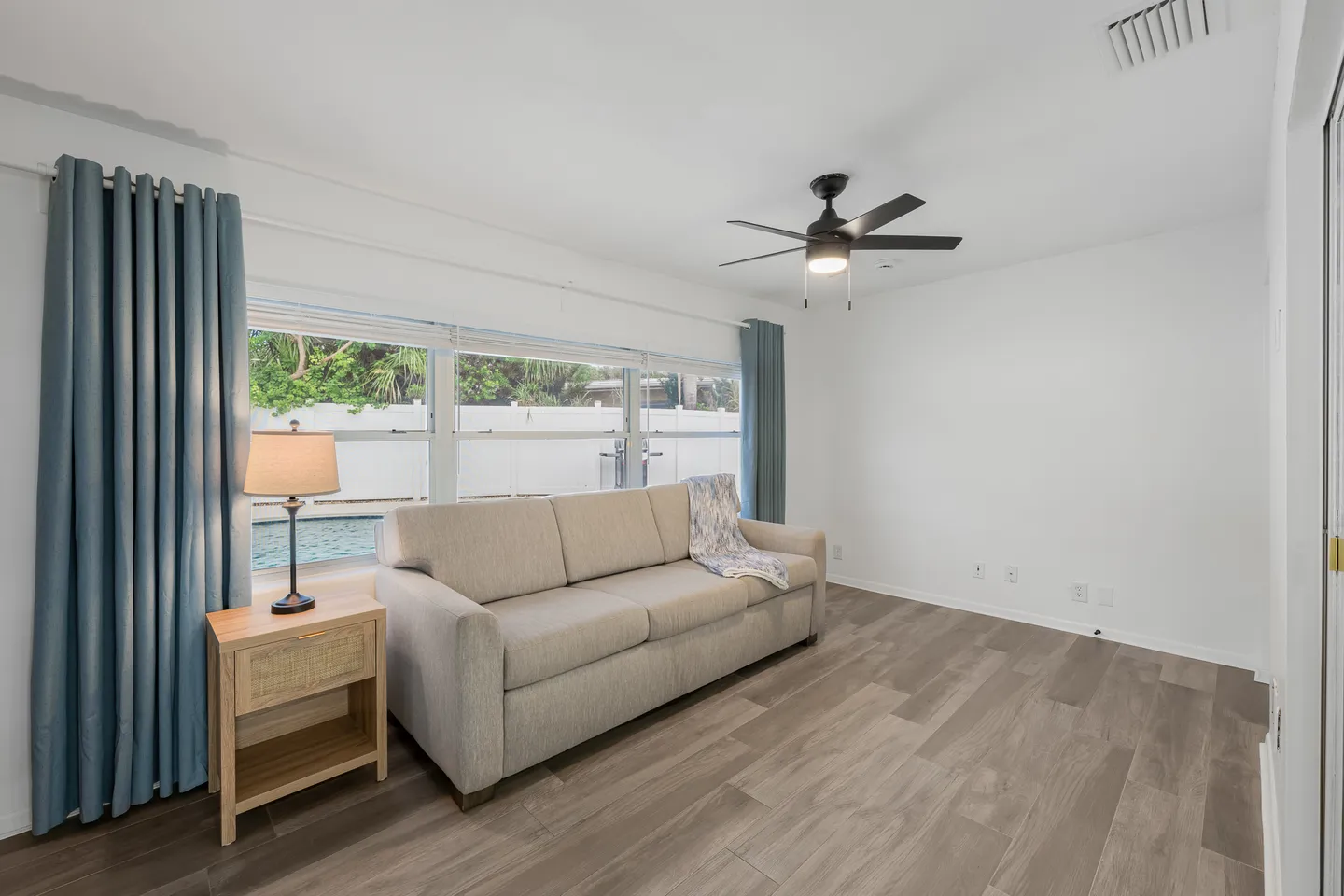 Living room featuring wood-look flooring, a beige sofa in front of large windows, blue curtains, a ceiling fan, and a view of an outdoor pool.