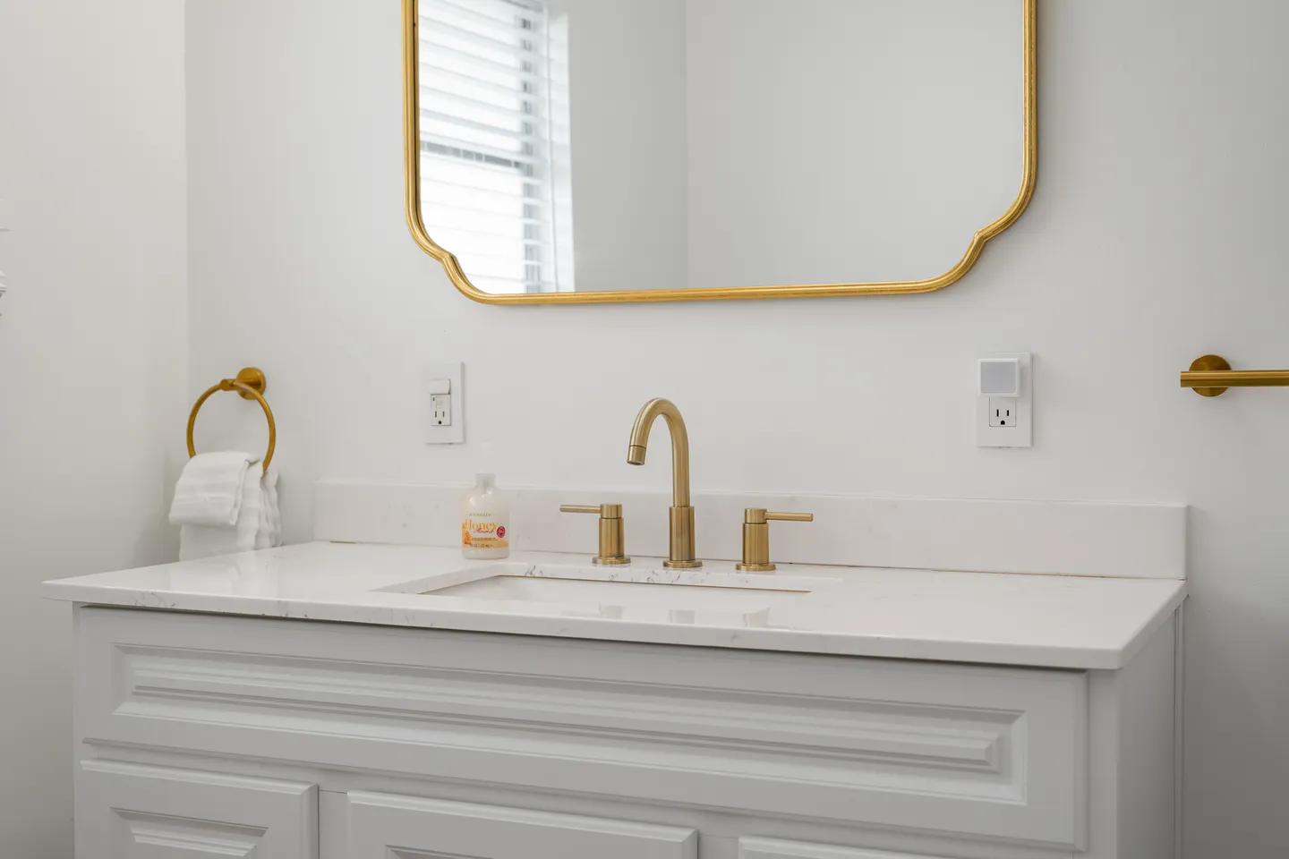A bathroom vanity with a white countertop, undermount sink, brass faucet, and a brass-framed mirror.