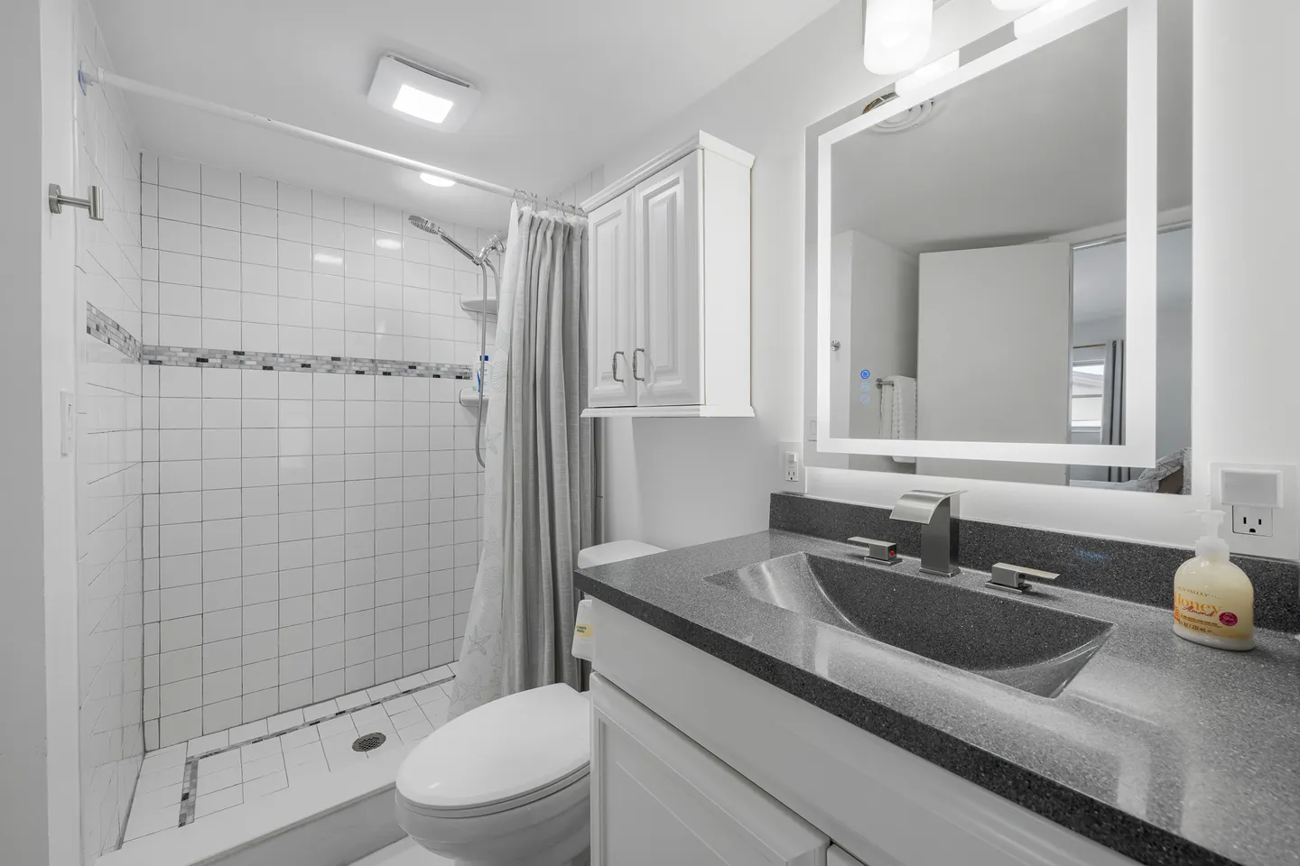 Bathroom with a dark countertop sink, lighted vanity mirror, toilet, and tiled walk-in shower with a curtain.