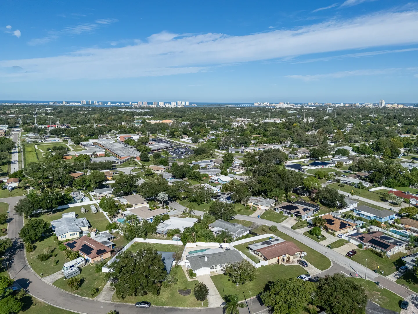 High aerial view of a suburban neighborhood showing single-story homes with yards, streets, trees, and the distant skyline under a blue sky.