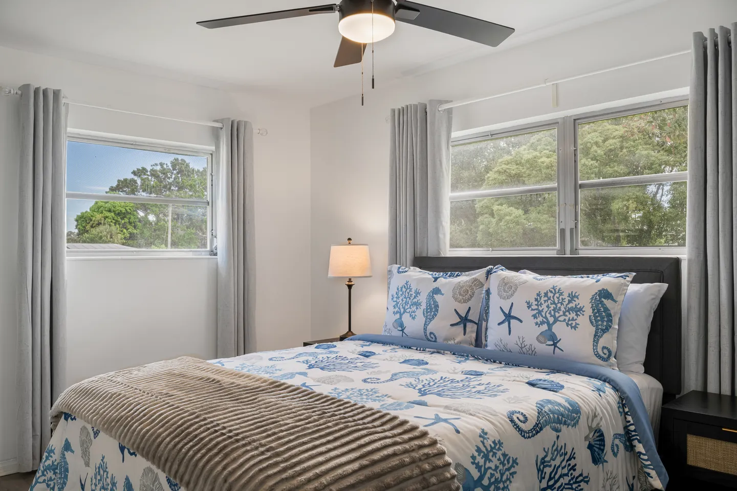 Bedroom featuring a made bed with blue and white coastal-patterned bedding, flanked by two windows and a nightstand with a lamp.