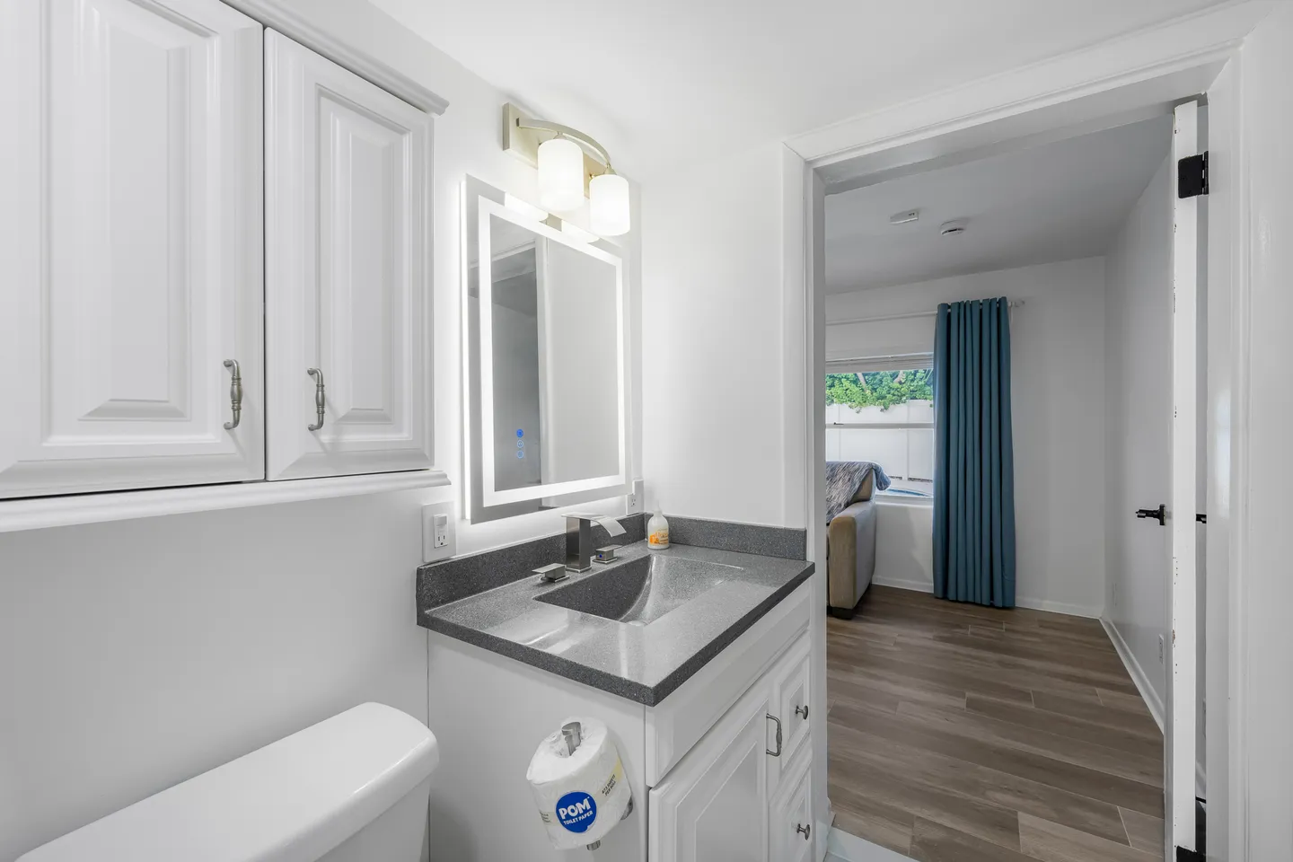 Bathroom with white cabinets, dark countertop, backlit vanity mirror, and view through an open doorway into an adjacent room.