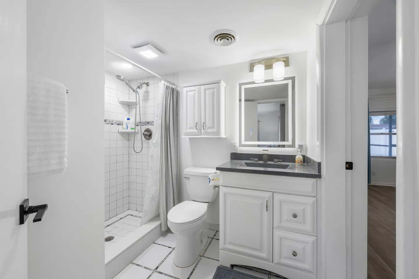 Bathroom with a walk-in tiled shower, white toilet, and single-sink vanity with dark countertop and lighted mirror.