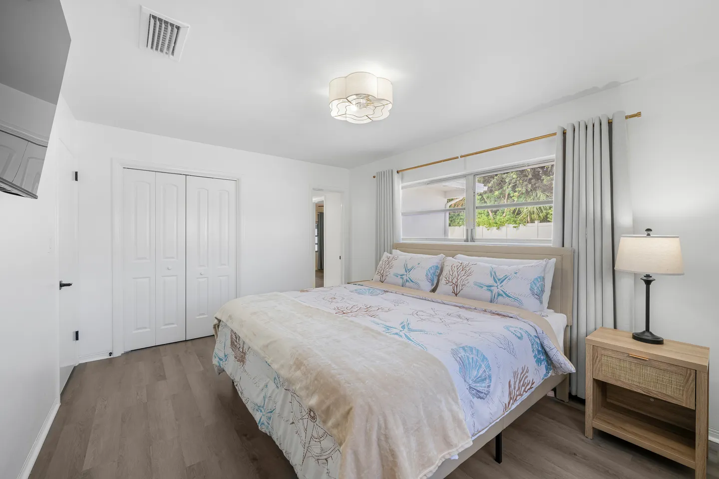 Bedroom featuring a made bed with coastal-themed bedding, a wooden nightstand with a lamp, a closet with bifold doors, and a window.