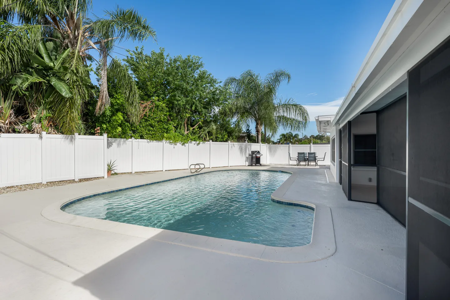Outdoor swimming pool in a private backyard surrounded by a white vinyl fence and lush green trees.