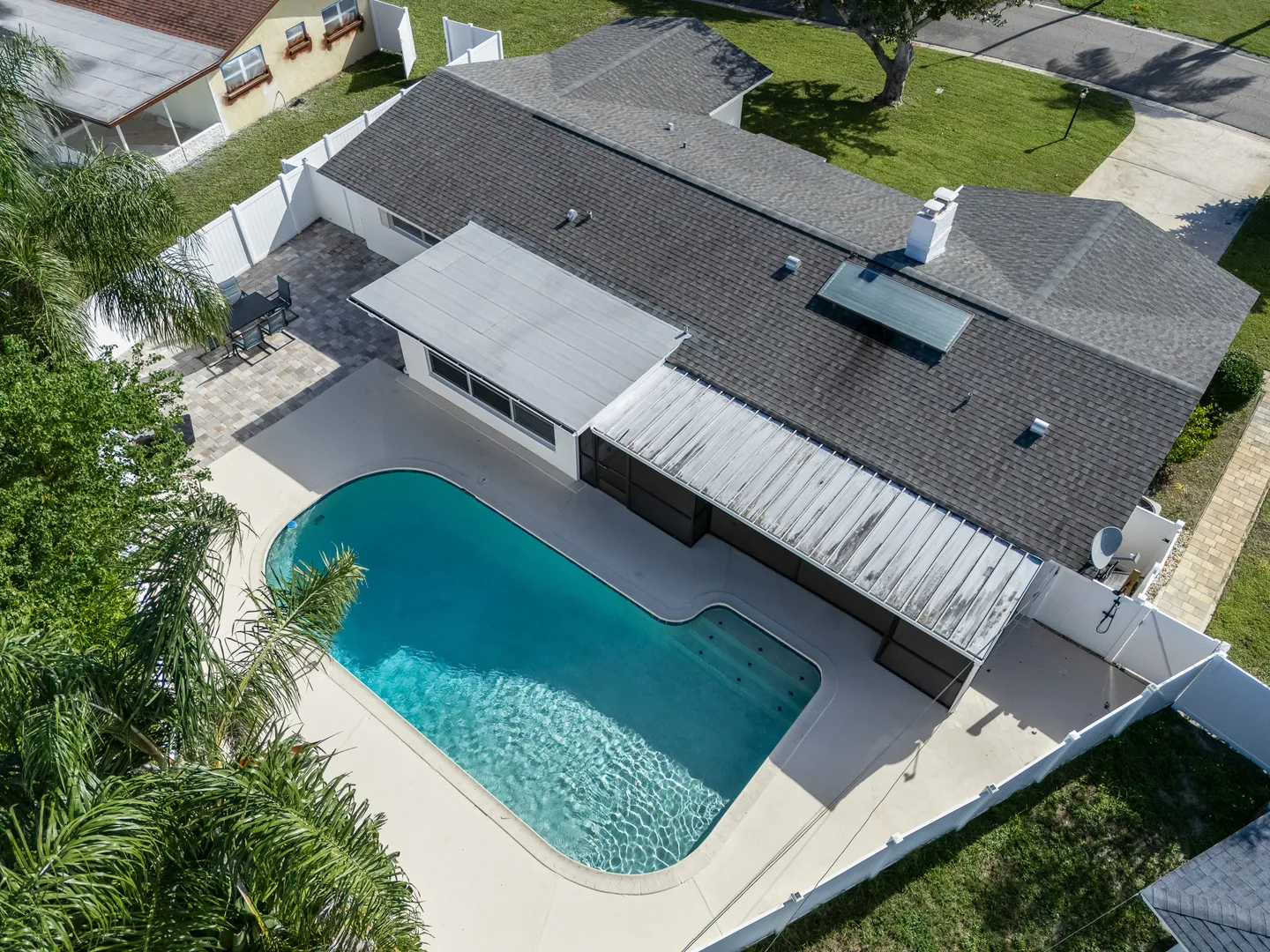 Aerial view of a backyard swimming pool, patio, and single-story home surrounded by a white fence.