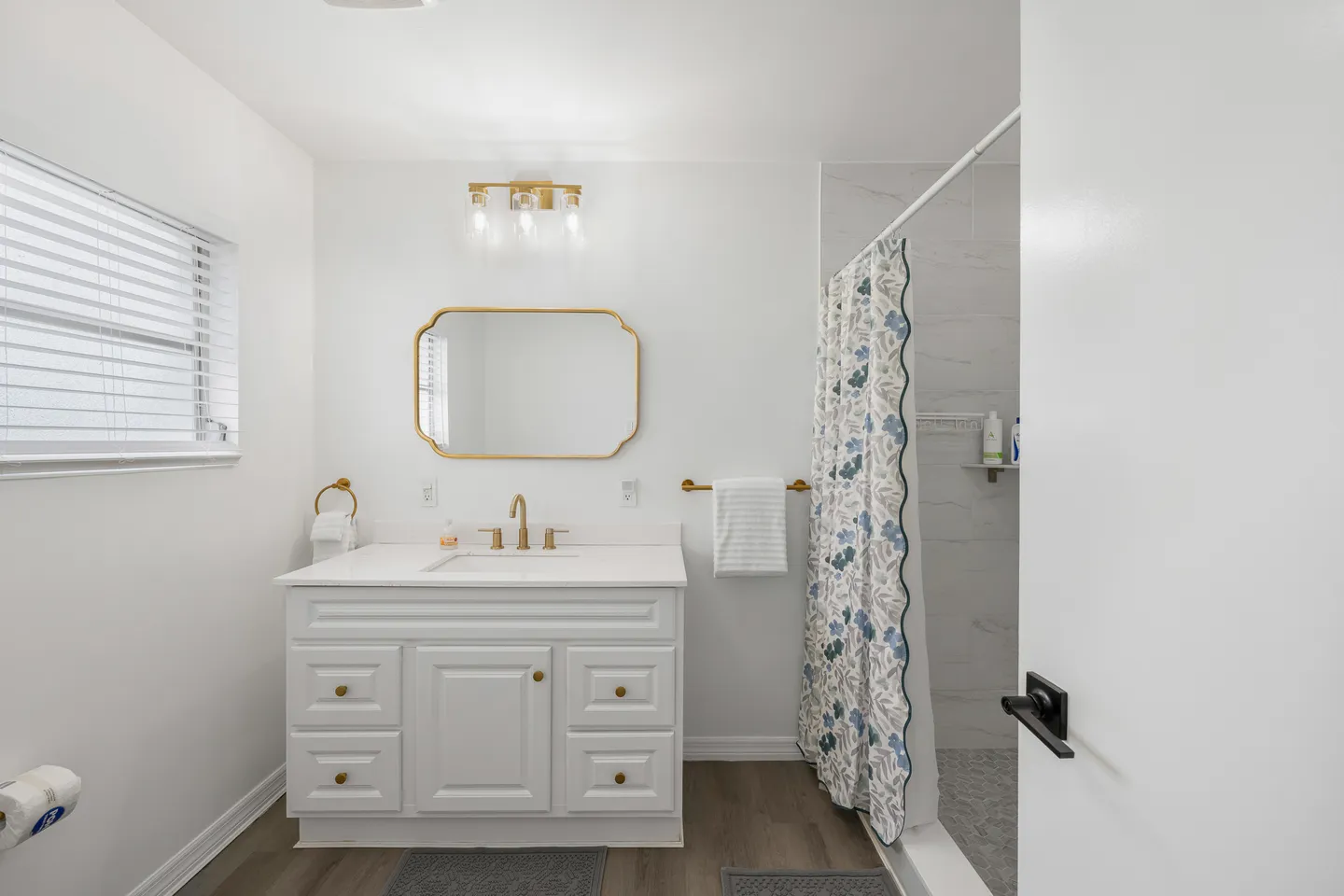 Bathroom with a white vanity, gold hardware, an arched mirror, and a walk-in shower with a floral curtain.