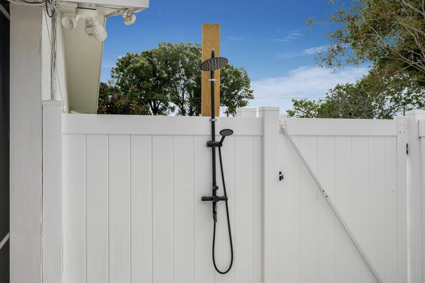 Outdoor shower with black fixtures mounted against a white vinyl privacy fence under a clear blue sky.