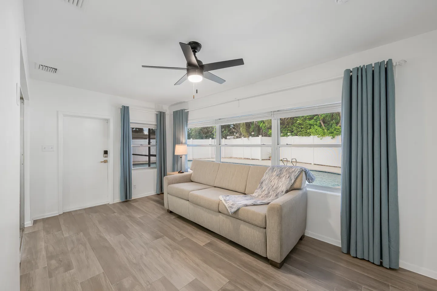 Bright room with wood-style flooring, a light-colored sofa, ceiling fan, and large windows looking out to a backyard pool.