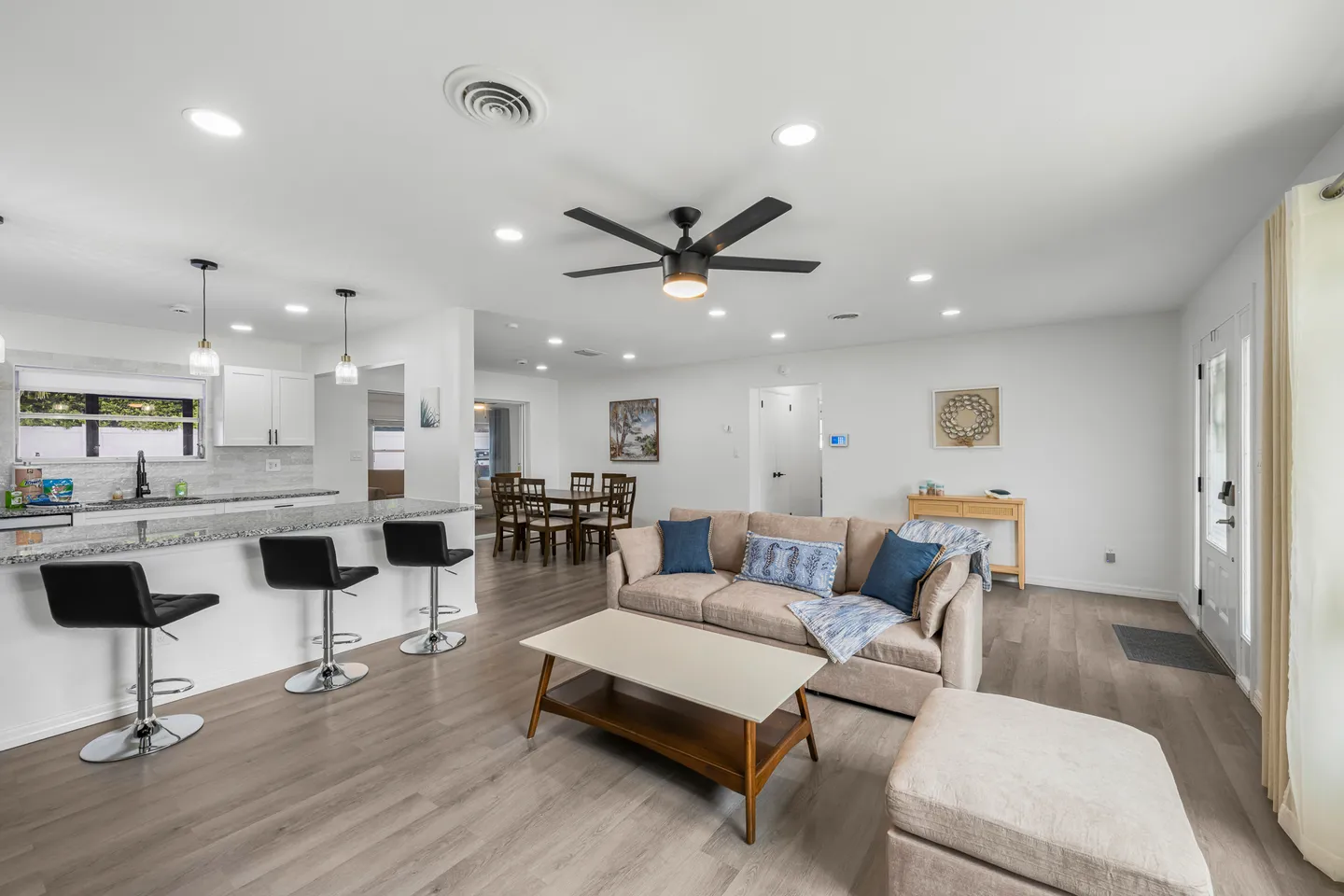 Open-concept living room with a tan couch and coffee table, adjacent to a kitchen counter with barstools and a dining area in the background.