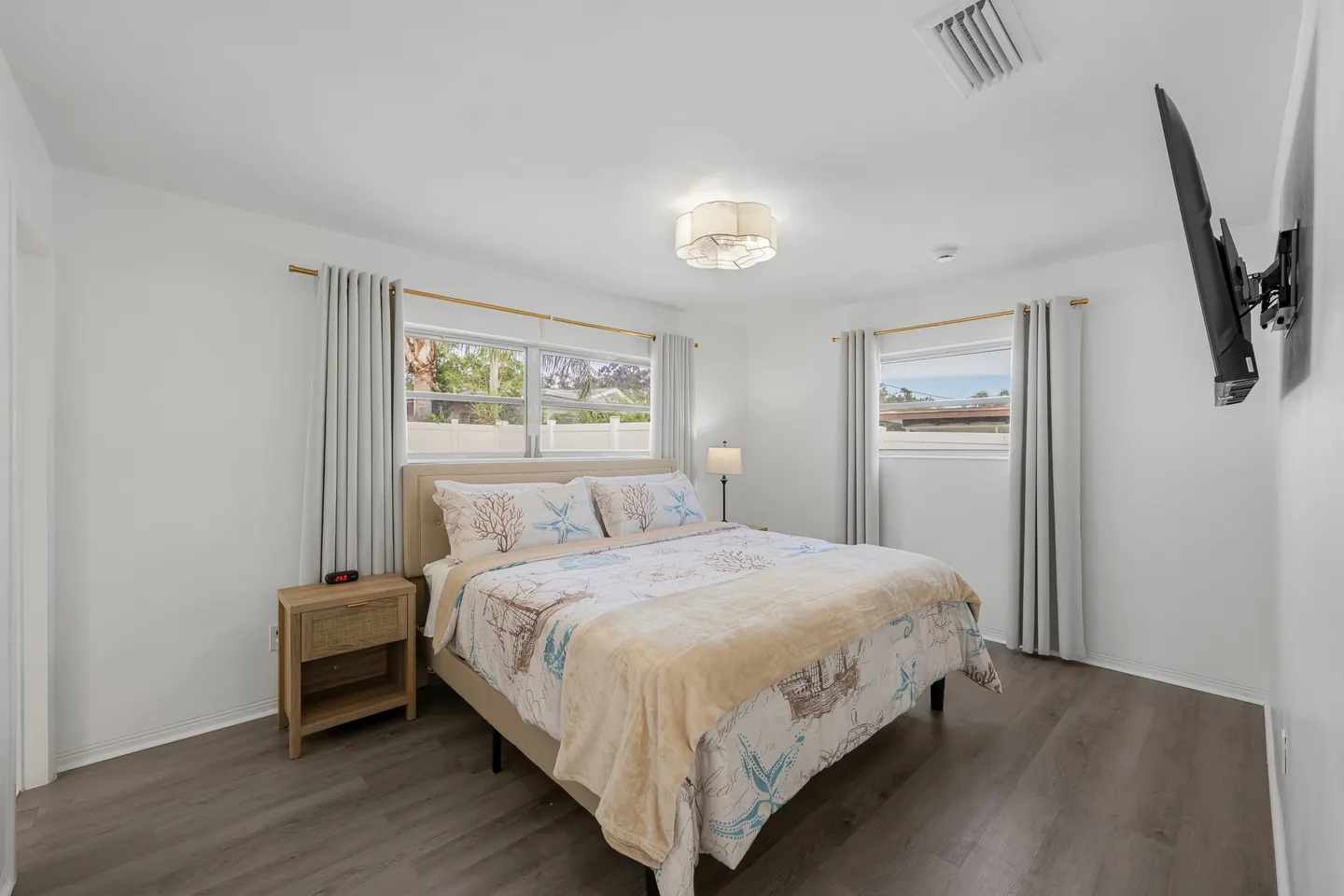 Bedroom with an upholstered bed, coastal-themed bedding, wood-look flooring, two curtained windows, and a wall-mounted TV.