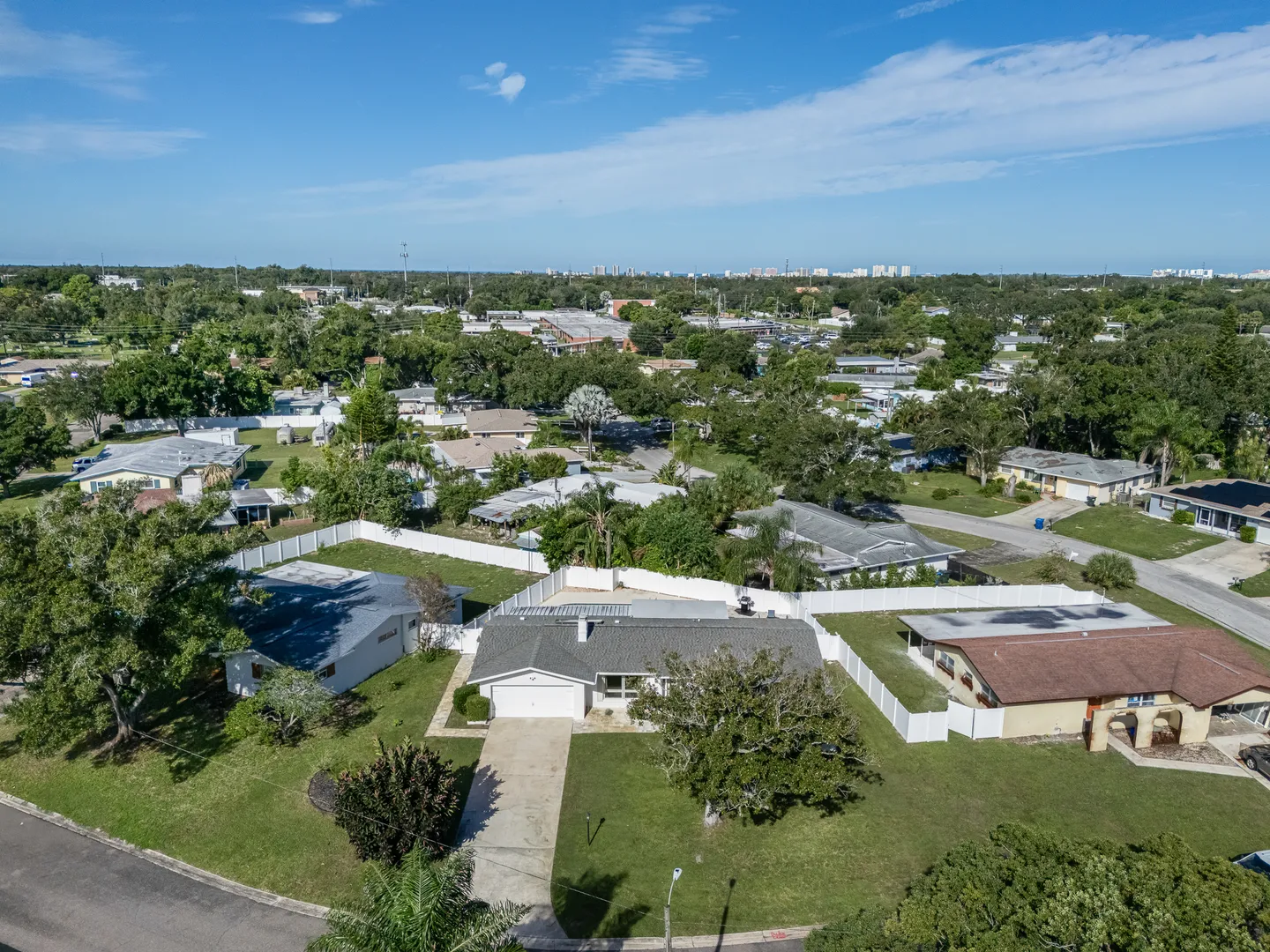 Aerial view of a single-story house with a long driveway, fenced yard, and surrounding residential neighborhood under a blue sky.