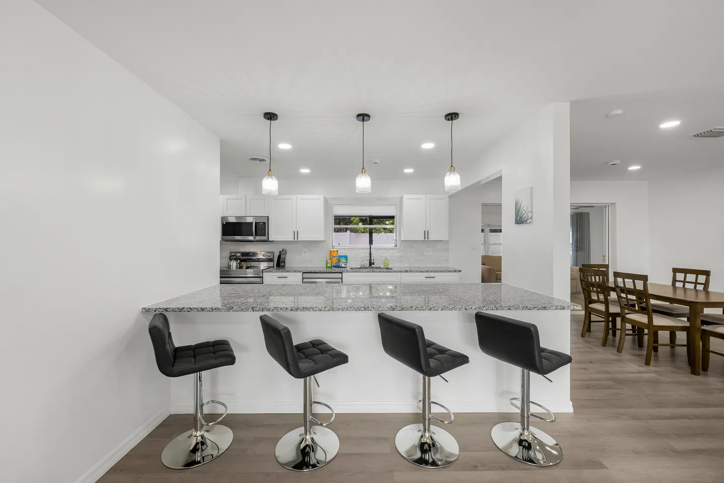 A kitchen breakfast bar with four black stools and three pendant lights, opening to the kitchen and dining area.