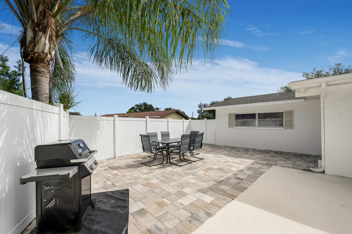 Outdoor patio with stone pavers, a gas barbecue grill, a patio dining set with six chairs, enclosed by a white vinyl fence under a palm tree and blue sky.